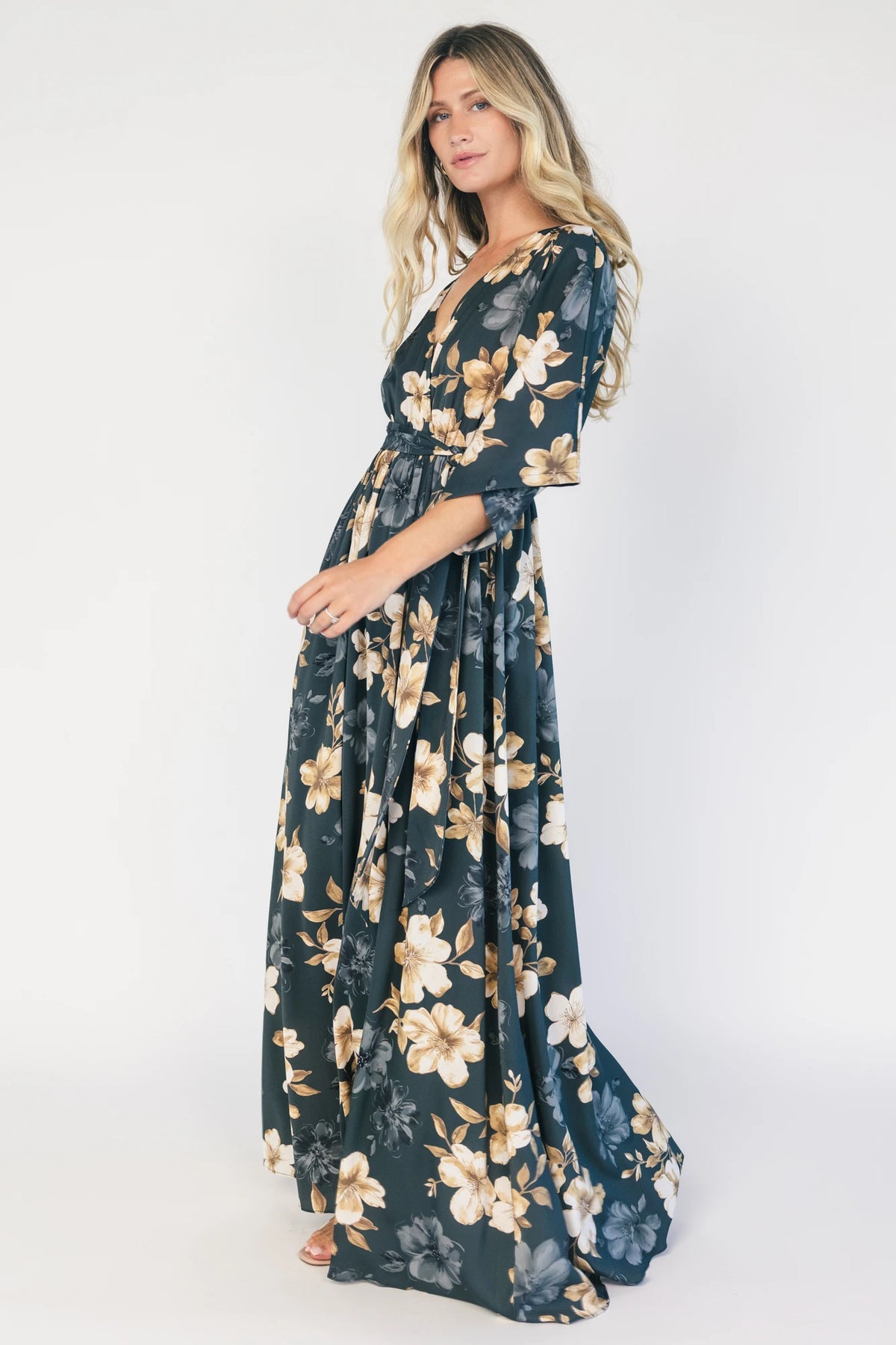 Model wearing a dark jade floral maxi dress with a surplice V-neckline, kimono sleeves, and an elastic waist with a removable sash tie.