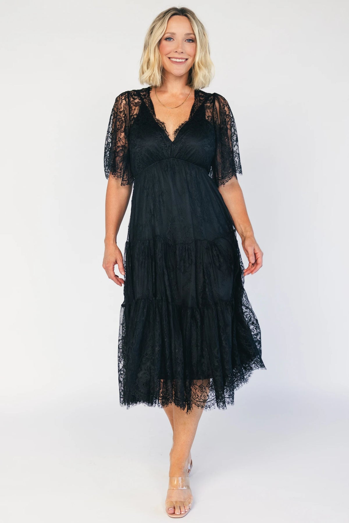 Model wearing a black lace midi dress with a deep V-neckline, short sheer scalloped sleeves, and an elastic waist.