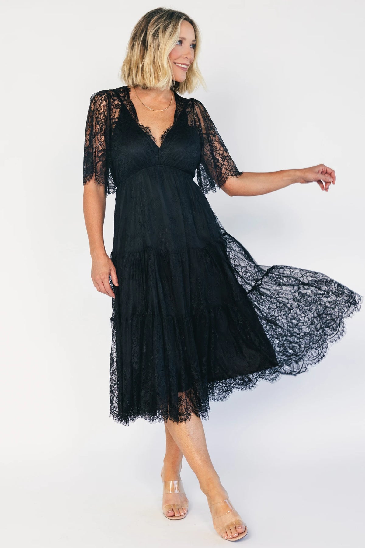 Model wearing a black lace midi dress with a deep V-neckline, short sheer scalloped sleeves, and an elastic waist.