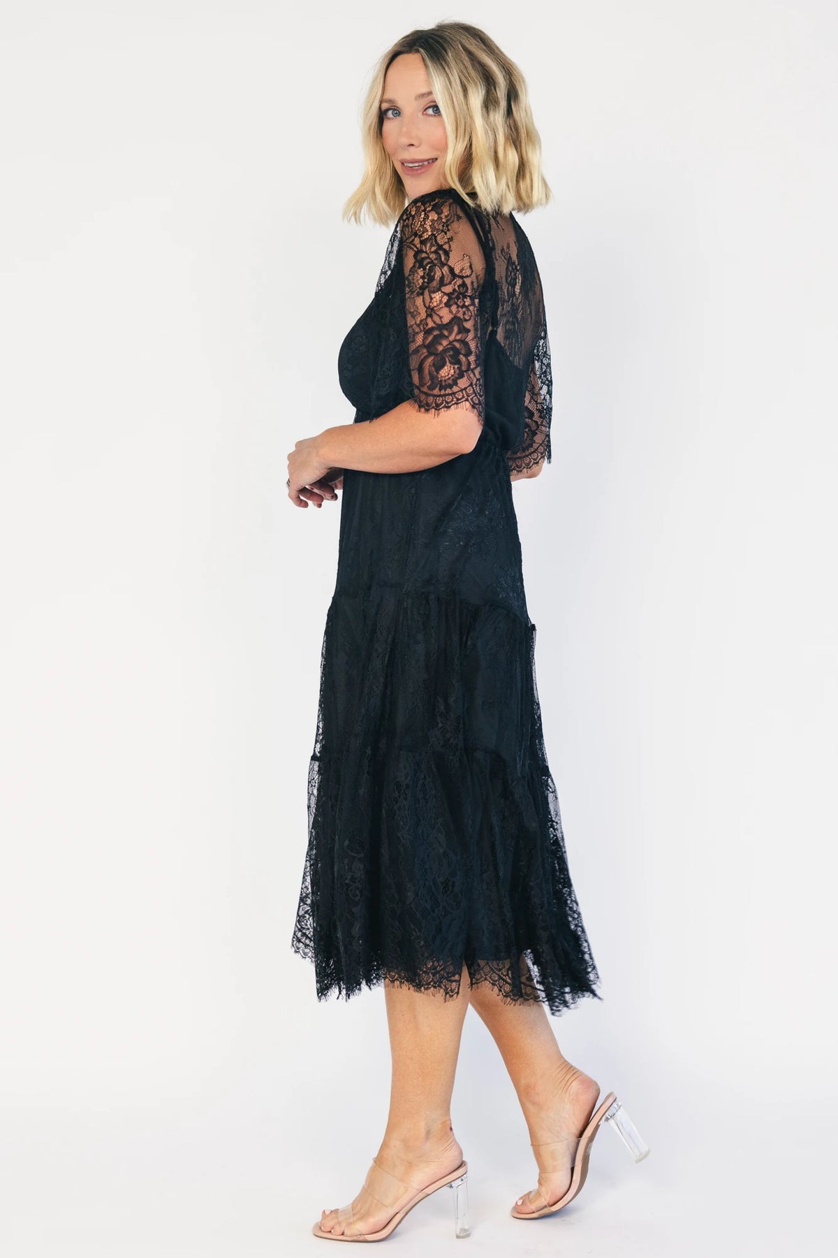 Model wearing a black lace midi dress with a deep V-neckline, short sheer scalloped sleeves, and an elastic waist.