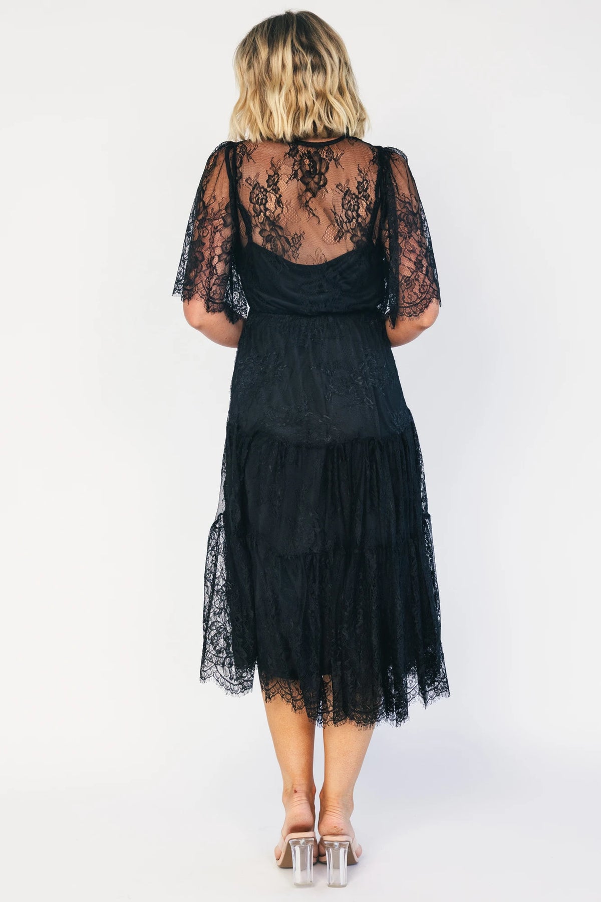 Model wearing a black lace midi dress with a deep V-neckline, short sheer scalloped sleeves, and an elastic waist.