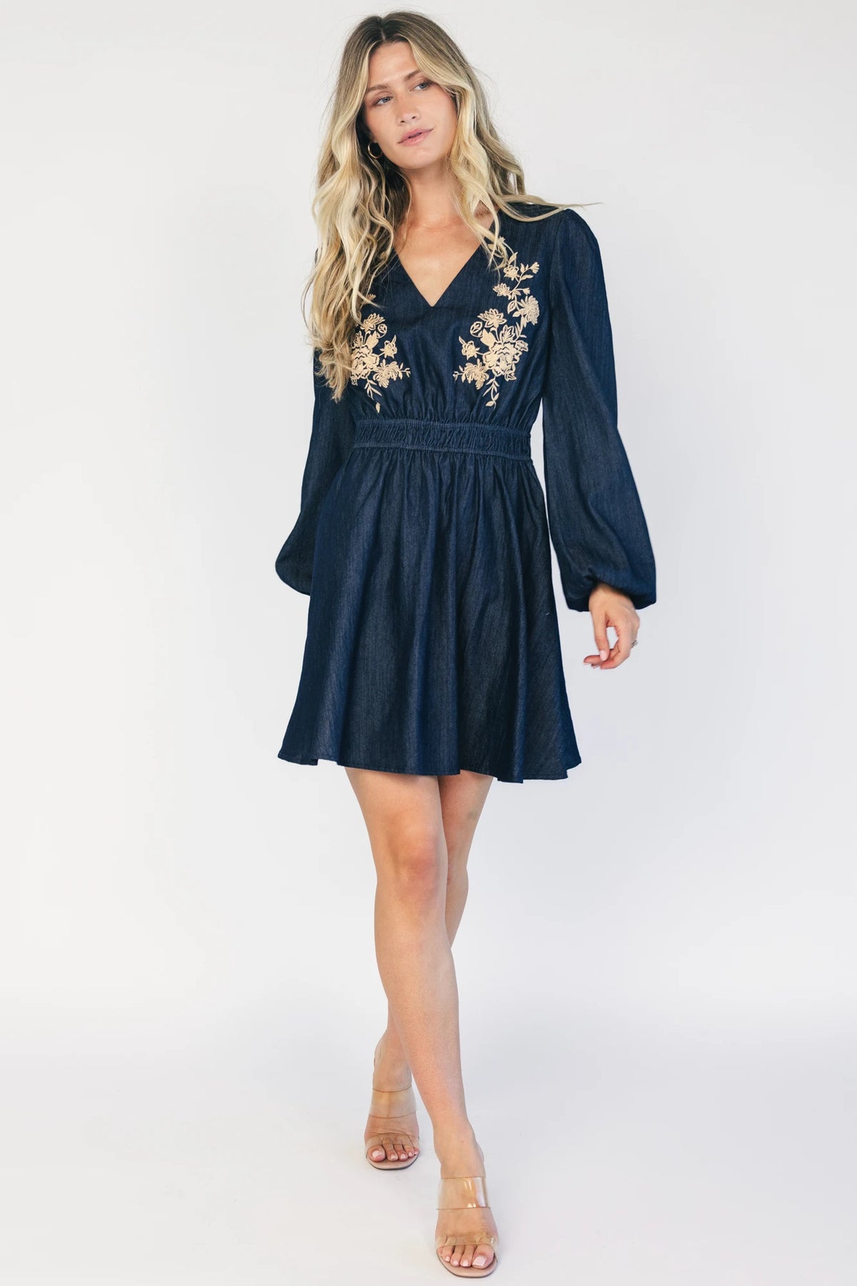 Model wearing a dark chambray short dress with a V-neckline, floral embroidery at the bust, long sleeves with elastic cuffs, and a double elastic waistband.