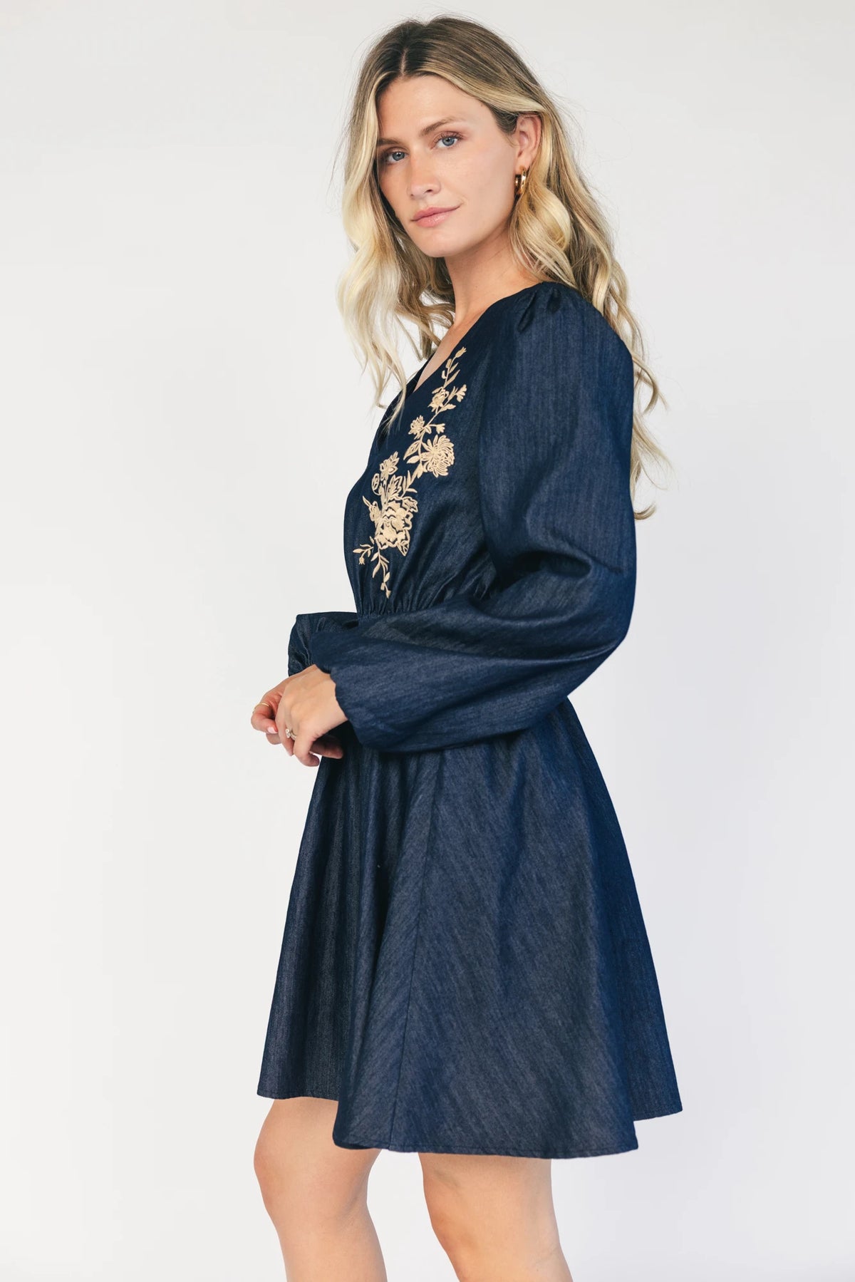 Model wearing a dark chambray short dress with a V-neckline, floral embroidery at the bust, long sleeves with elastic cuffs, and a double elastic waistband.
