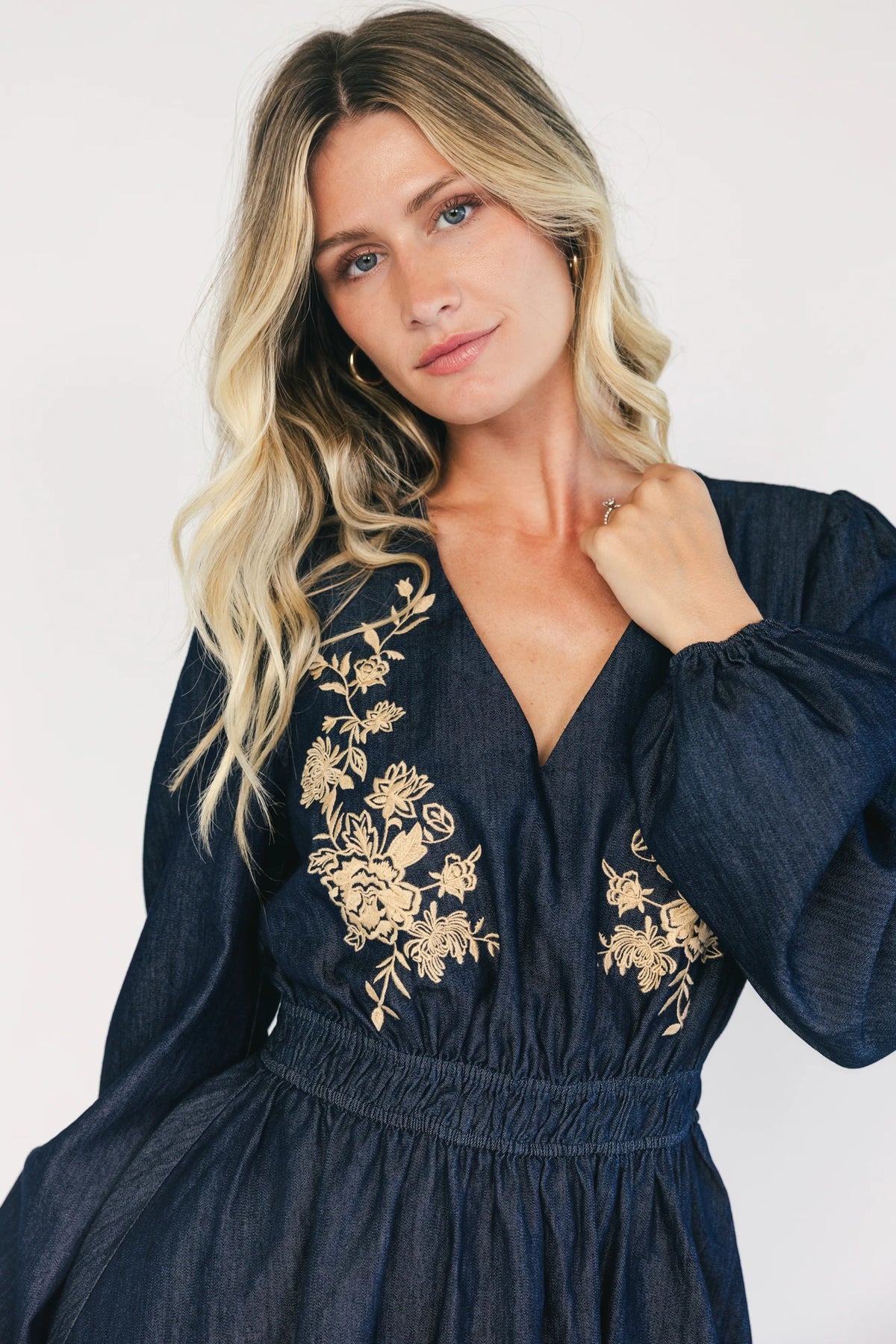 Model wearing a dark chambray short dress with a V-neckline, floral embroidery at the bust, long sleeves with elastic cuffs, and a double elastic waistband.