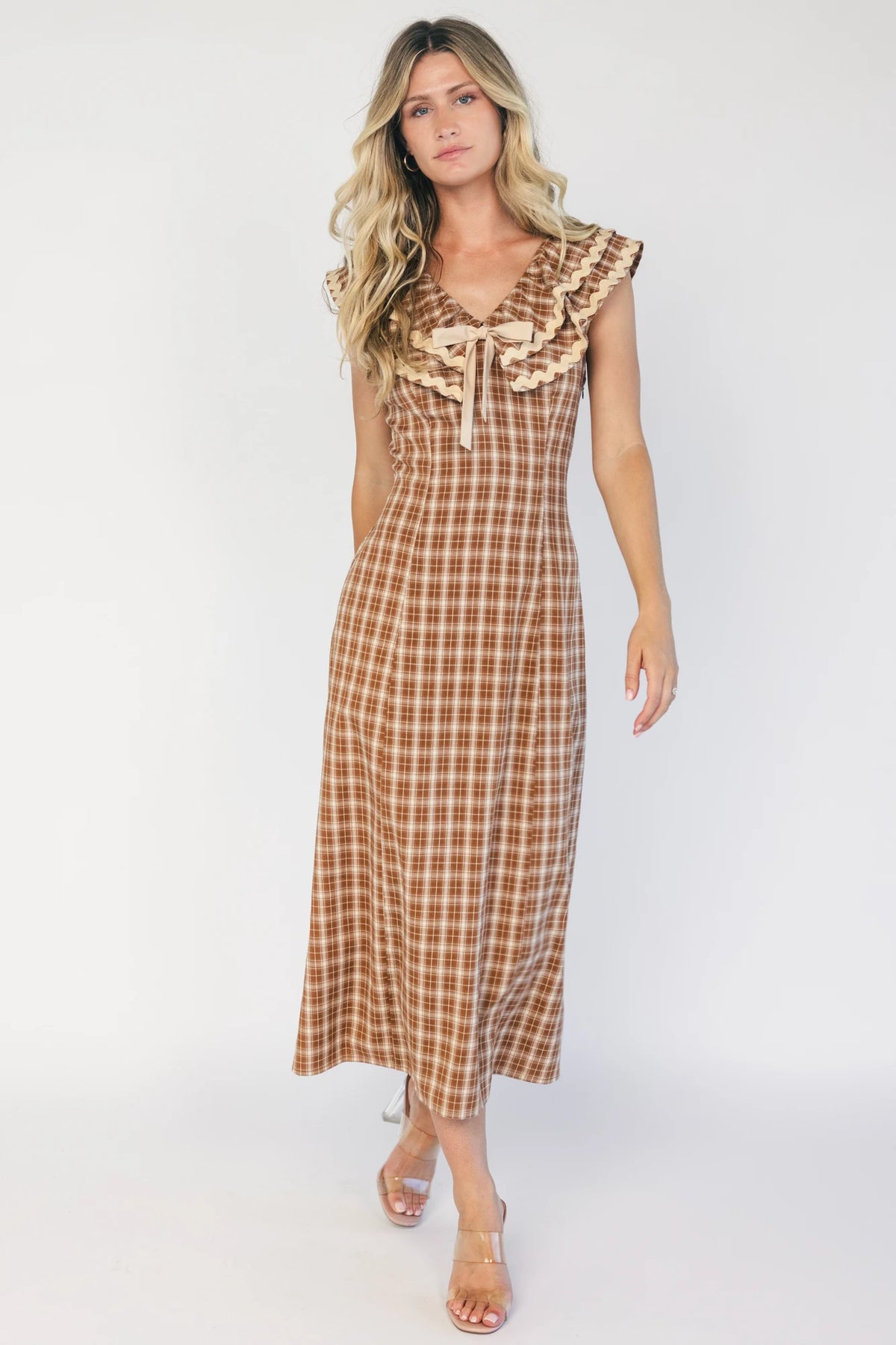 Model wearing a dusty mocha and cream plaid midi dress with a V-neckline, decorative bow tie detail, cascading ruffle trim, and a fitted silhouette.