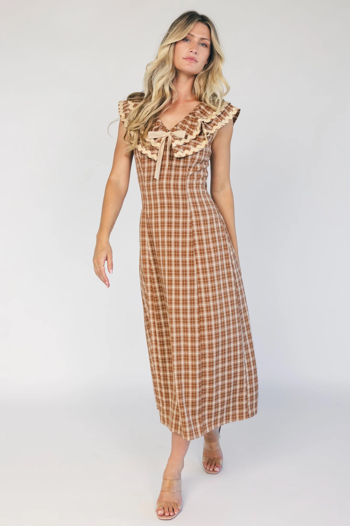 Model wearing a dusty mocha and cream plaid midi dress with a V-neckline, decorative bow tie detail, cascading ruffle trim, and a fitted silhouette.