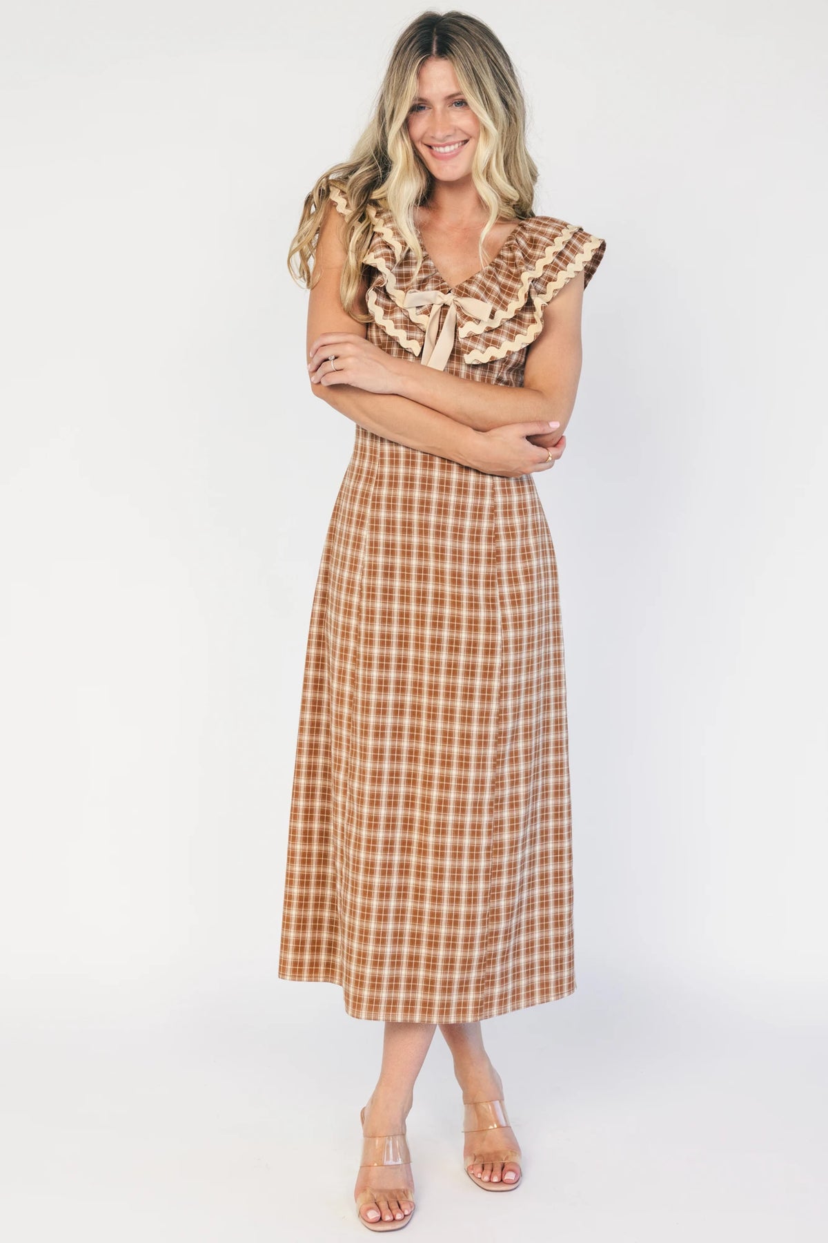 Model wearing a dusty mocha and cream plaid midi dress with a V-neckline, decorative bow tie detail, cascading ruffle trim, and a fitted silhouette.
