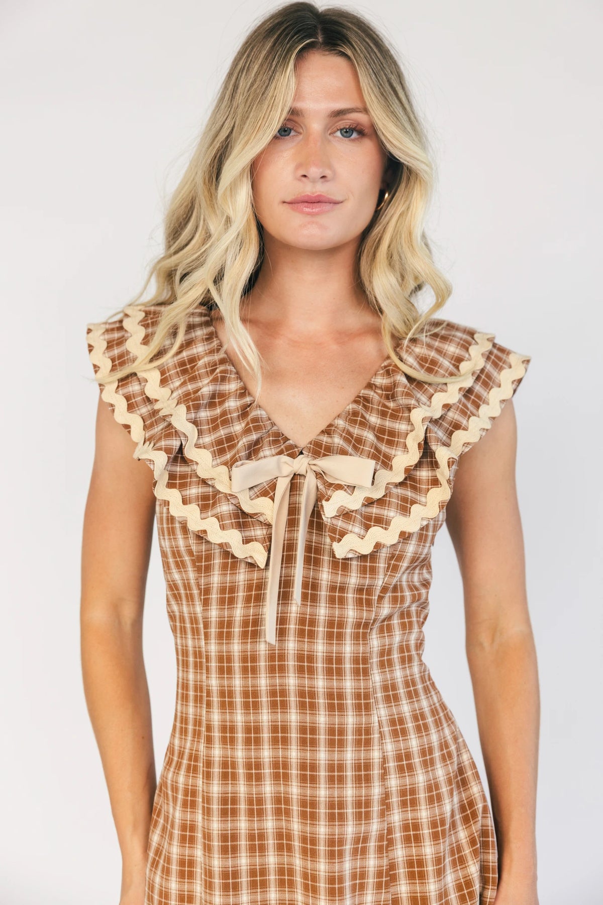 Model wearing a dusty mocha and cream plaid midi dress with a V-neckline, decorative bow tie detail, cascading ruffle trim, and a fitted silhouette.