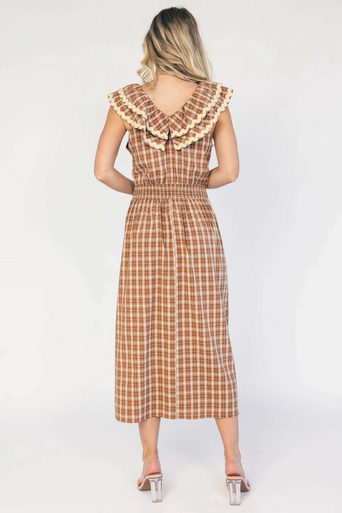 Model wearing a dusty mocha and cream plaid midi dress with a V-neckline, decorative bow tie detail, cascading ruffle trim, and a fitted silhouette.
