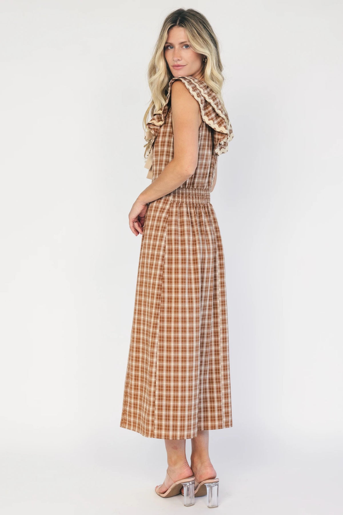 Model wearing a dusty mocha and cream plaid midi dress with a V-neckline, decorative bow tie detail, cascading ruffle trim, and a fitted silhouette.