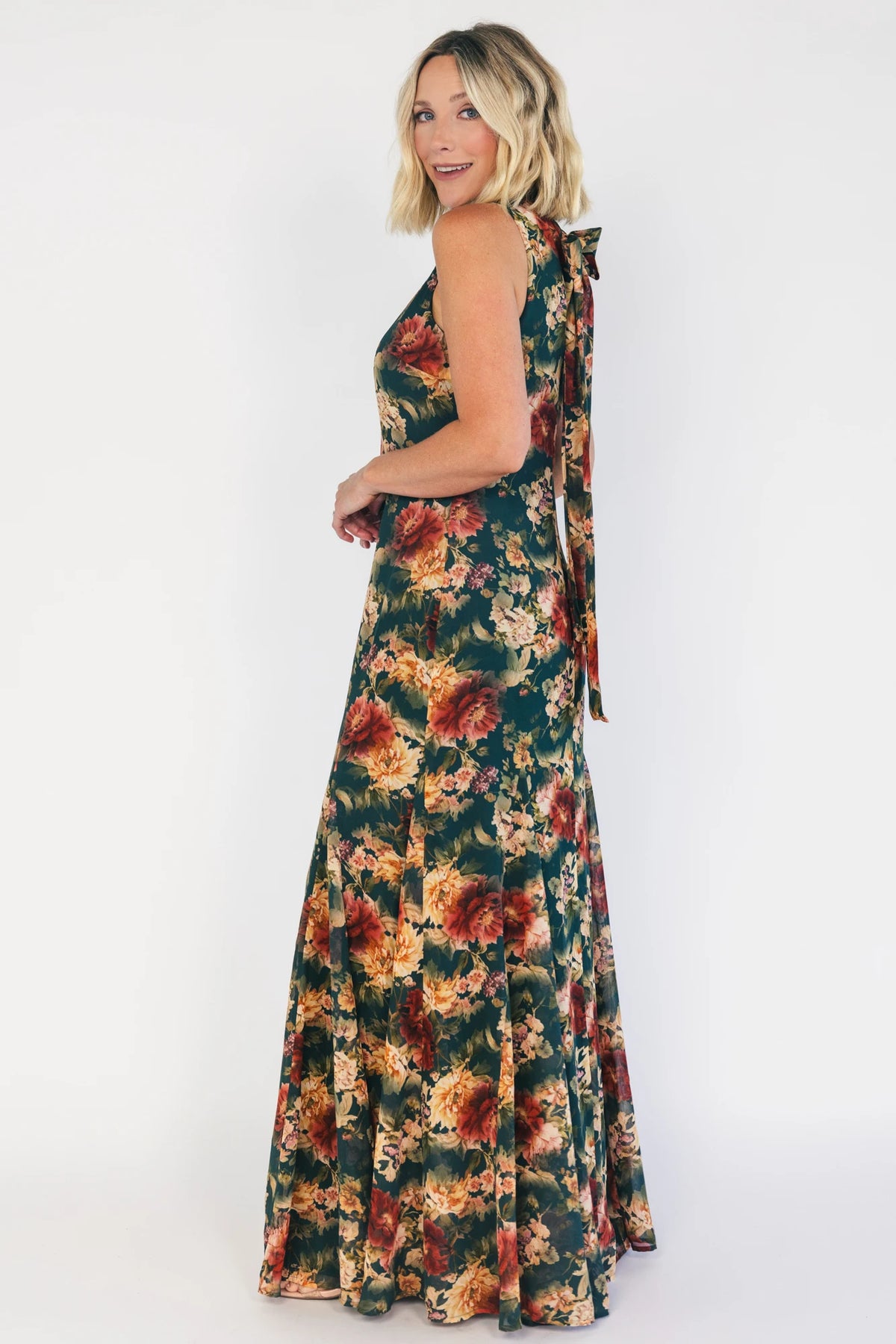 Model wearing a topaz floral maxi dress with a high round neckline, sleeveless bodice, and an open back with a sash tie.