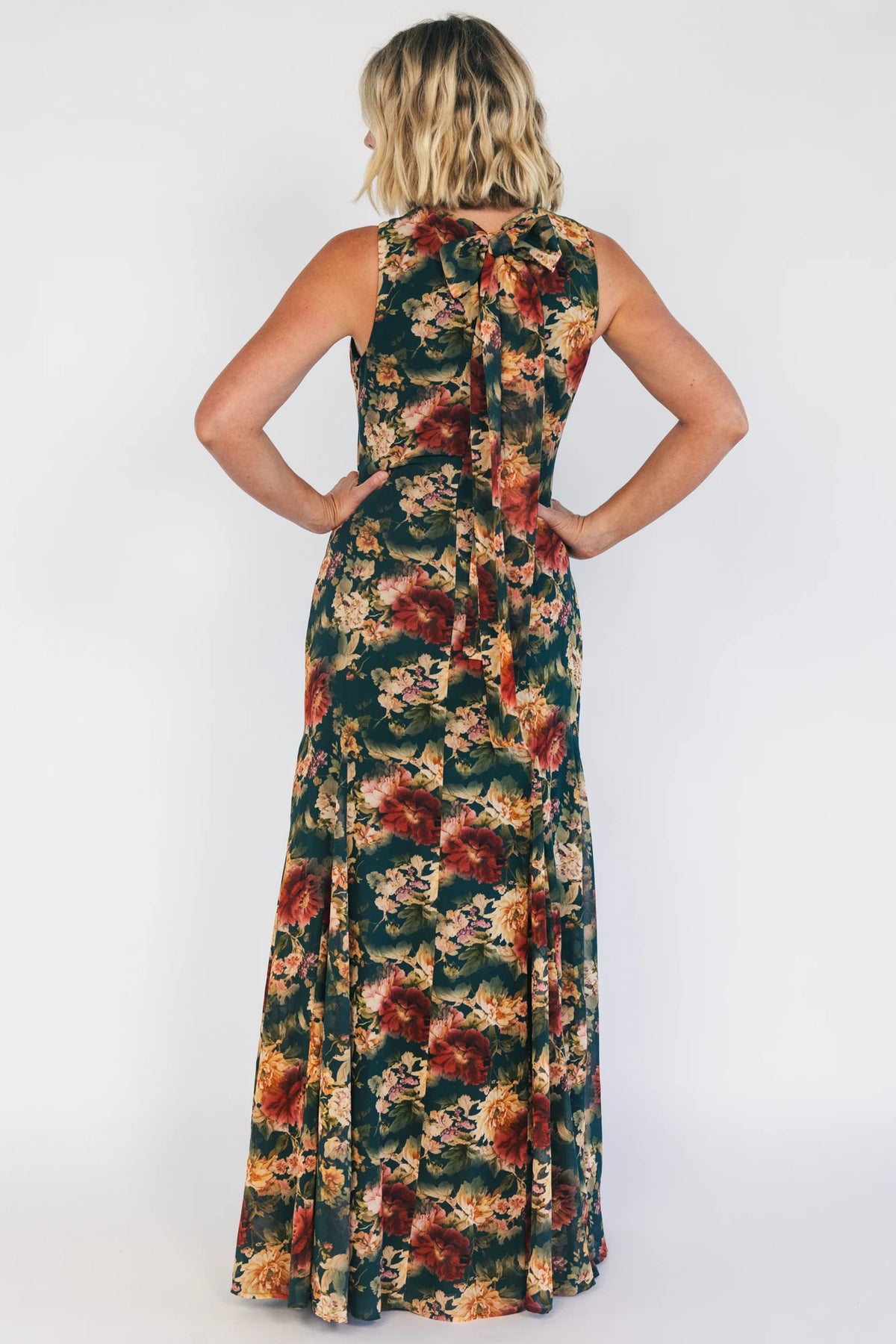 Model wearing a topaz floral maxi dress with a high round neckline, sleeveless bodice, and an open back with a sash tie.