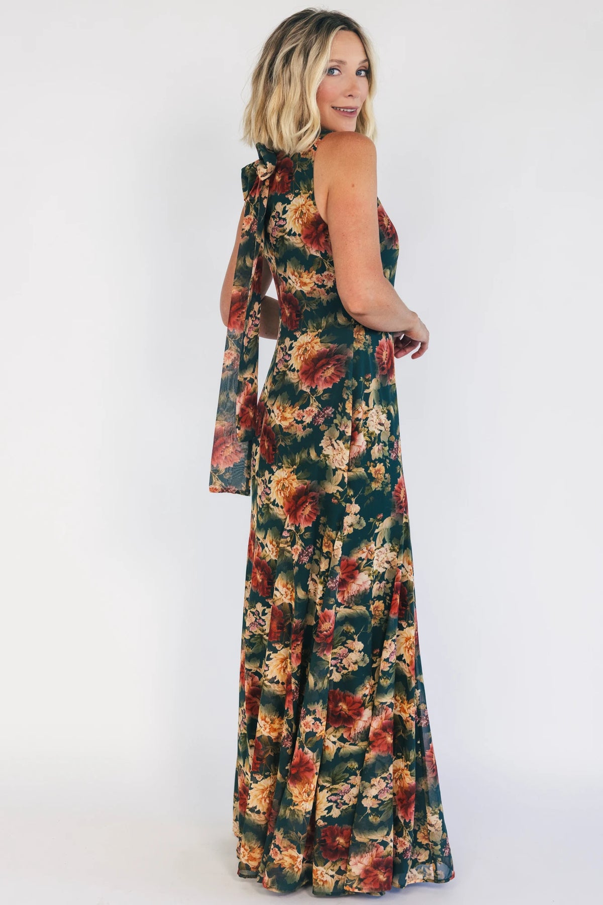 Model wearing a topaz floral maxi dress with a high round neckline, sleeveless bodice, and an open back with a sash tie.