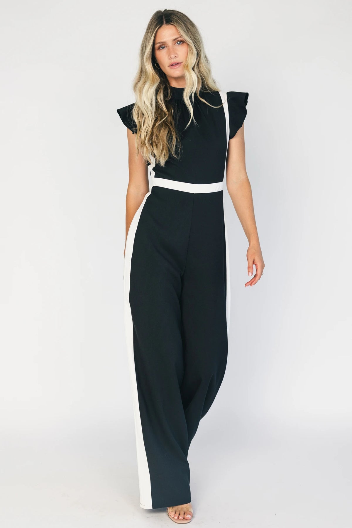 Model wearing the Lumina Ruffle Jumpsuit in Black + Ivory featuring a sleeveless ruffle-trim bodice, defined waist, and flowing wide-leg pants.