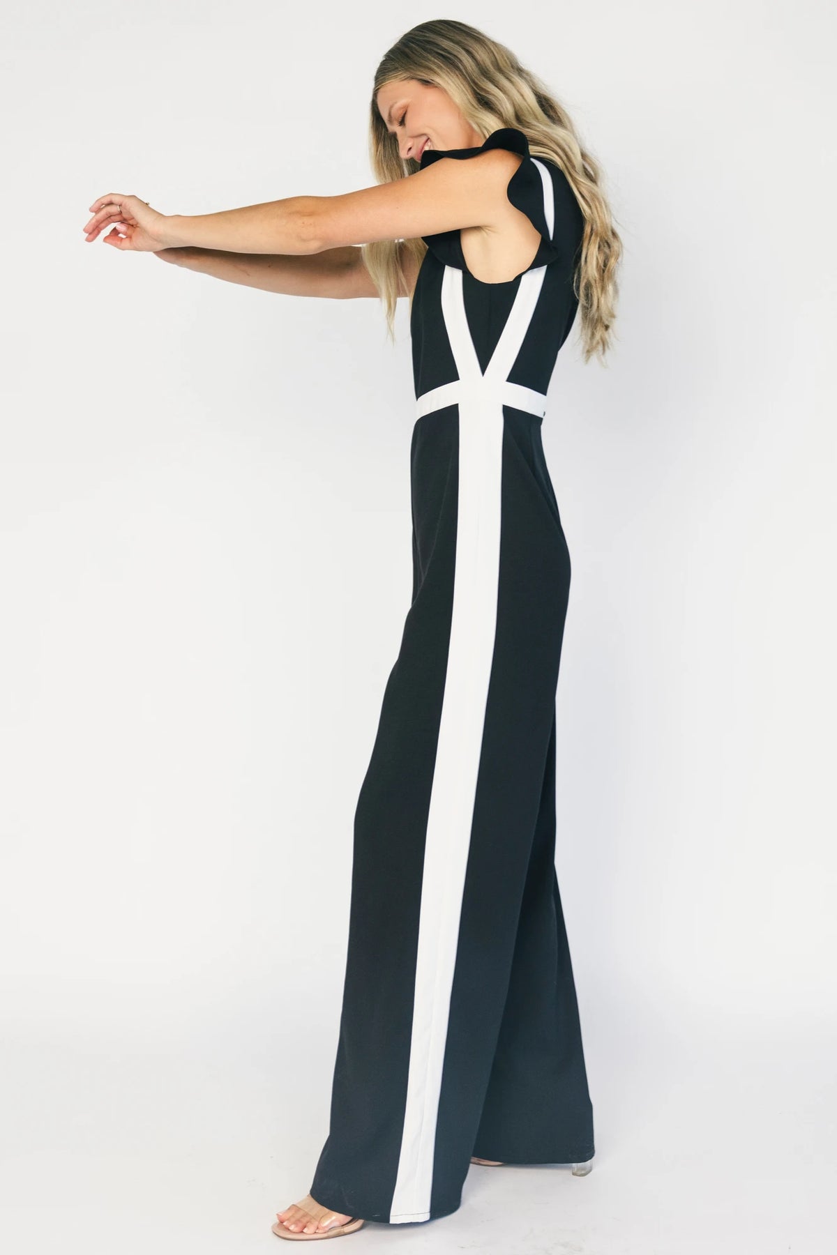 Model wearing the Lumina Ruffle Jumpsuit in Black + Ivory featuring a sleeveless ruffle-trim bodice, defined waist, and flowing wide-leg pants.