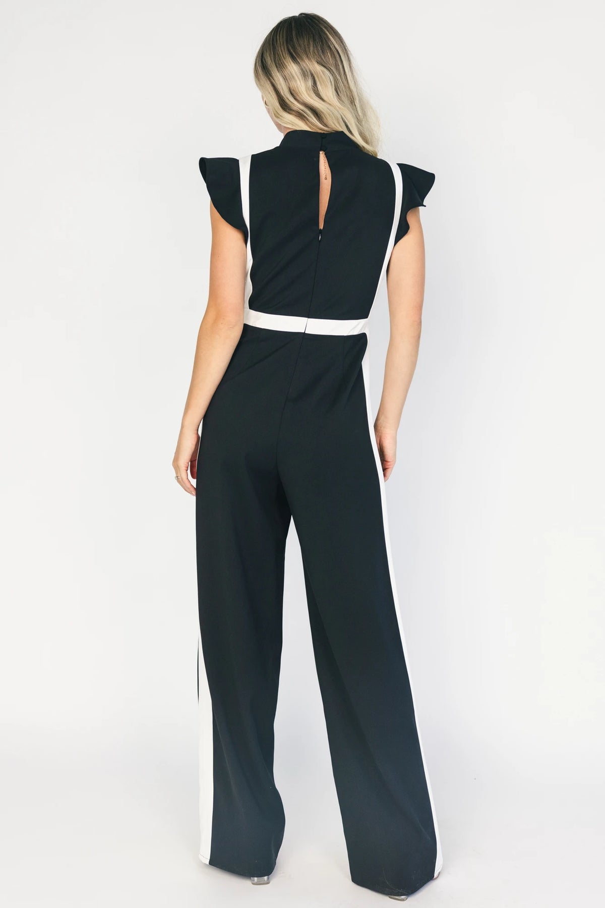 Model wearing the Lumina Ruffle Jumpsuit in Black + Ivory featuring a sleeveless ruffle-trim bodice, defined waist, and flowing wide-leg pants.
