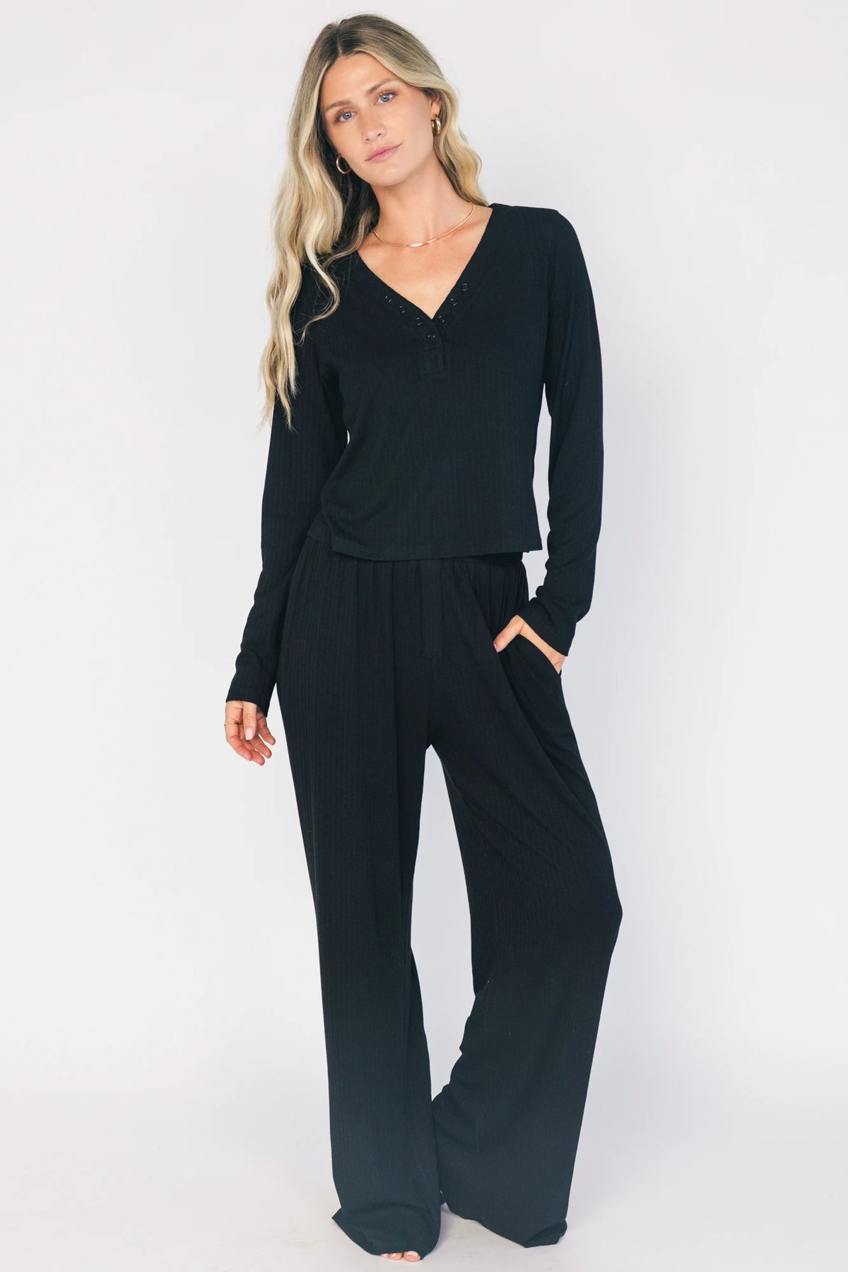 Model wearing the Rubi Soft Ribbed Pajama Set in Black featuring a soft ribbed knit, long-sleeve V-neck top with a snap-button placket, and matching straight-leg pants with an elastic waistband and side pockets.