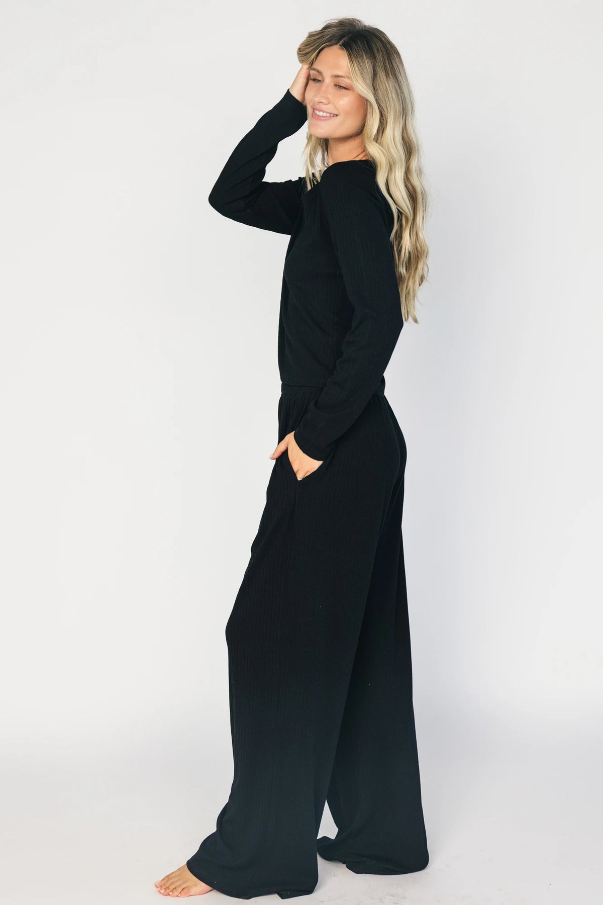 Model wearing the Rubi Soft Ribbed Pajama Set in Black featuring a soft ribbed knit, long-sleeve V-neck top with a snap-button placket, and matching straight-leg pants with an elastic waistband and side pockets.