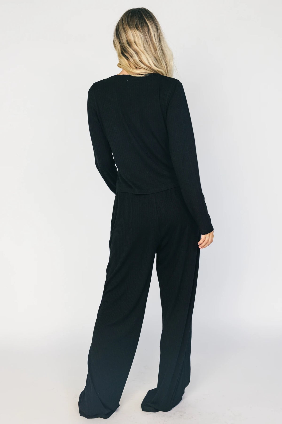 Model wearing the Rubi Soft Ribbed Pajama Set in Black featuring a soft ribbed knit, long-sleeve V-neck top with a snap-button placket, and matching straight-leg pants with an elastic waistband and side pockets.