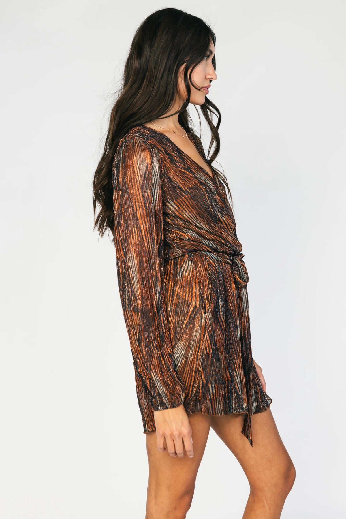 Model wearing the Nightfall Romper in Bronze Shimmer featuring a surplice V-neckline, long sleeves, an elastic waist with a removable sash tie, and flowy scalloped shorts in a metallic pleated shimmer fabric.