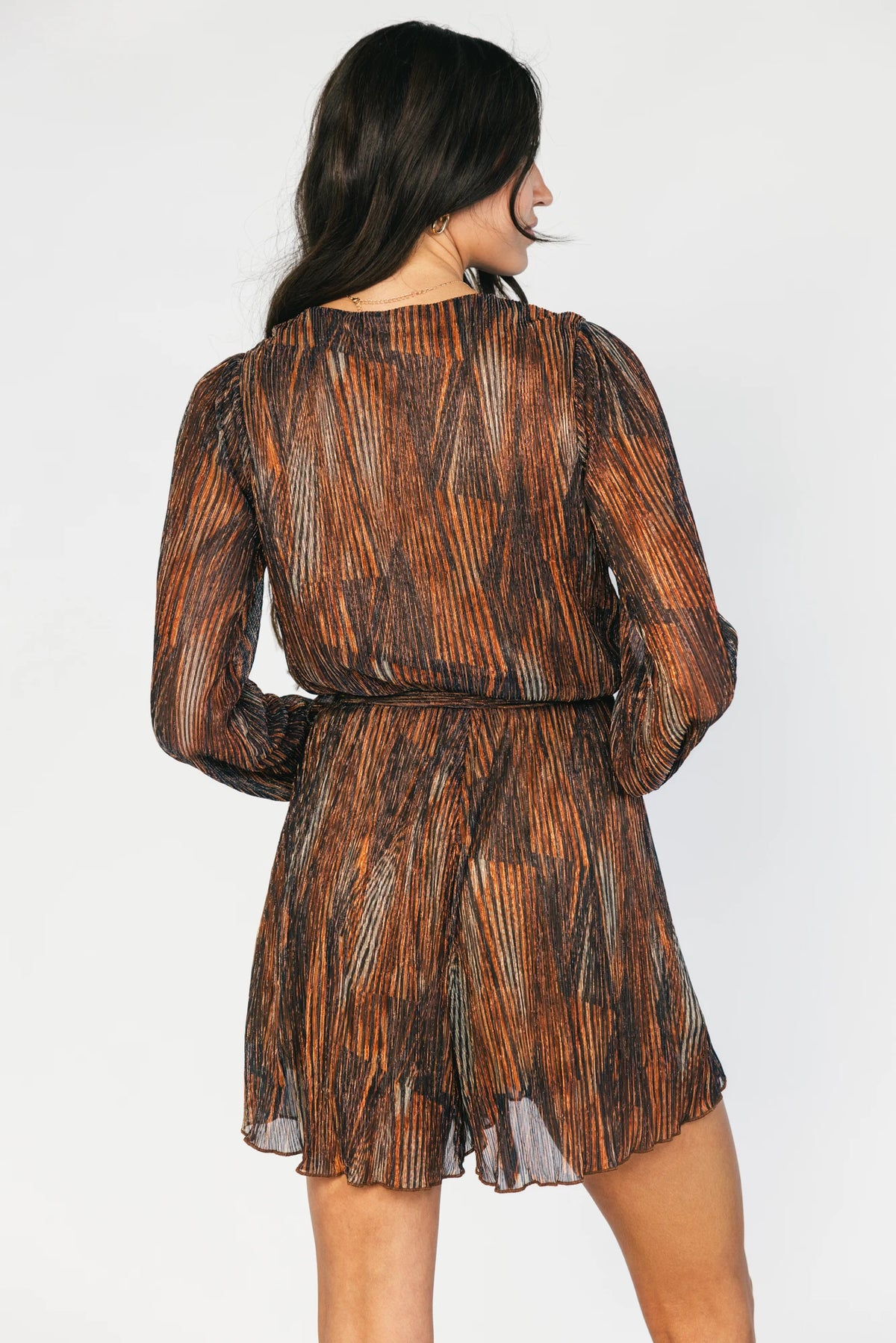Model wearing the Nightfall Romper in Bronze Shimmer featuring a surplice V-neckline, long sleeves, an elastic waist with a removable sash tie, and flowy scalloped shorts in a metallic pleated shimmer fabric.