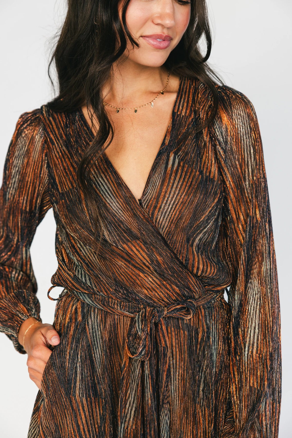 Model wearing the Nightfall Romper in Bronze Shimmer featuring a surplice V-neckline, long sleeves, an elastic waist with a removable sash tie, and flowy scalloped shorts in a metallic pleated shimmer fabric.