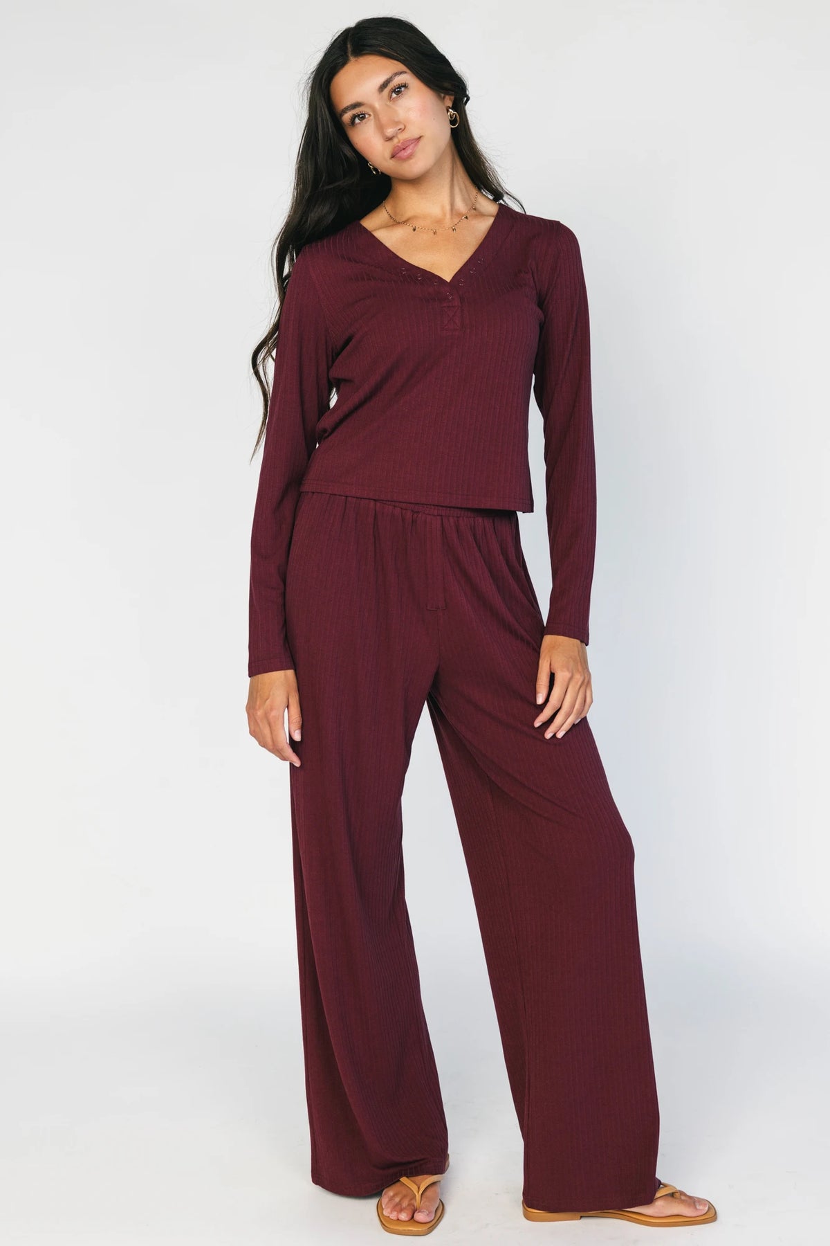 Model wearing the Rubi Soft Ribbed Pajama Set in Burgundy featuring a soft ribbed knit, long-sleeve V-neck top with a snap-button placket, and matching straight-leg pants with an elastic waistband and side pockets.