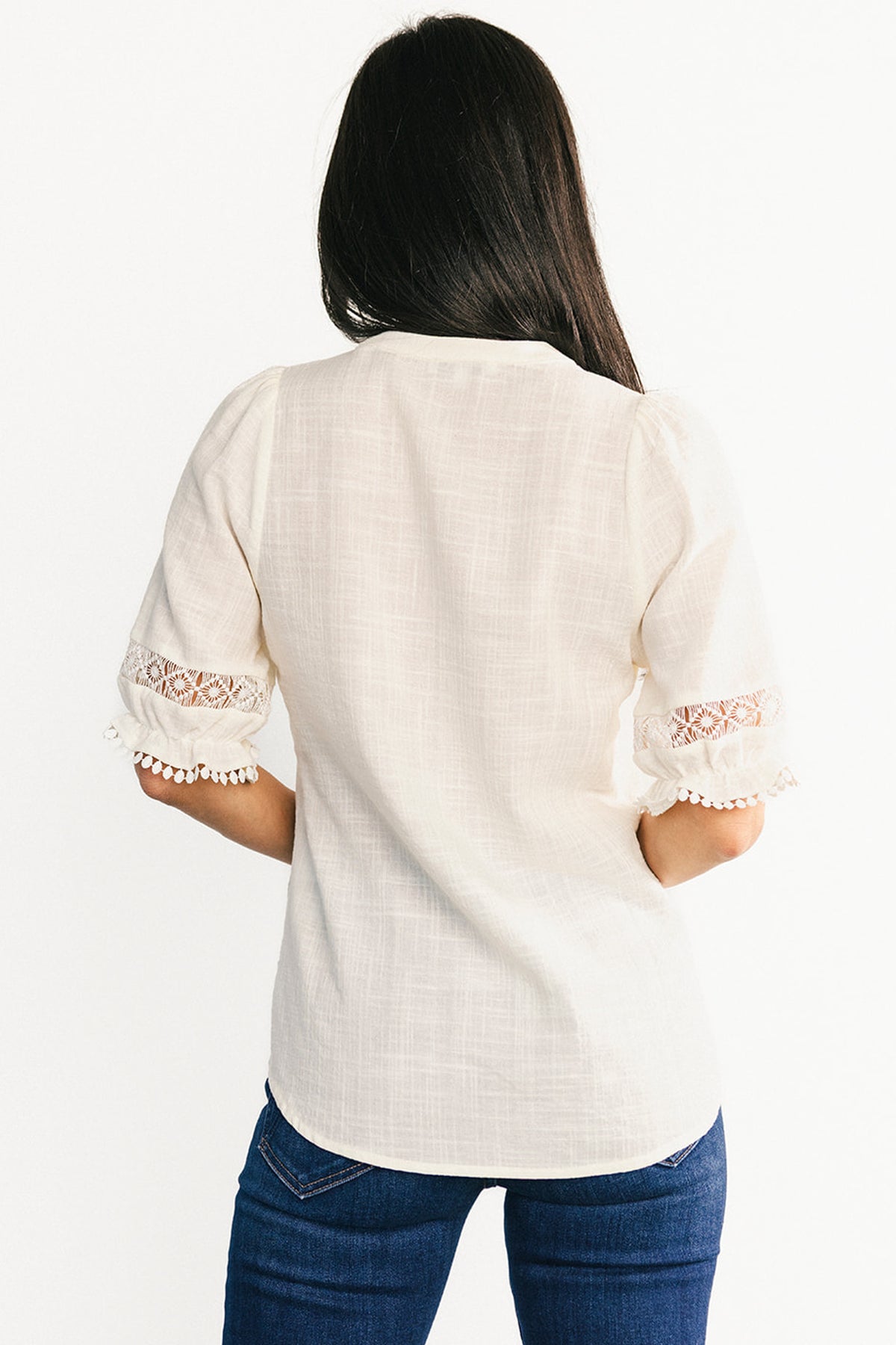 Textile close-up displaying the cotton-viscose linen-look weave, embroidered lace panel, and pom-pom cuff trim that define the Alba top's tactile details.
