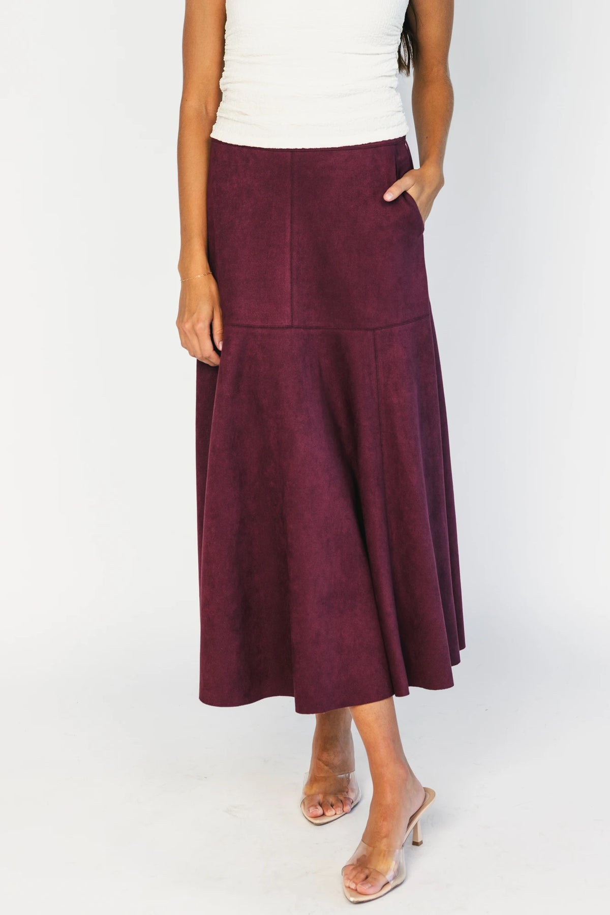 Model wearing a mulberry faux suede A-line midi skirt with a high-rise waistband, stitched panel detailing, side pockets, and a raw hem for a timeless, feminine silhouette.
