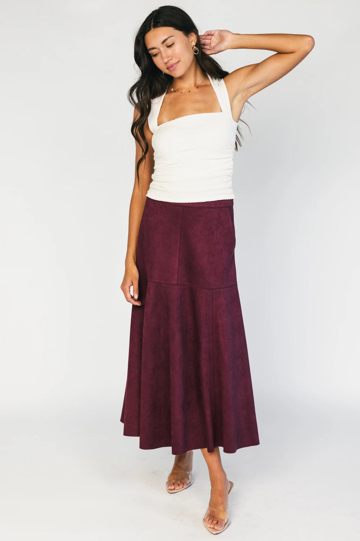 Model wearing a mulberry faux suede A-line midi skirt with a high-rise waistband, stitched panel detailing, side pockets, and a raw hem for a timeless, feminine silhouette.