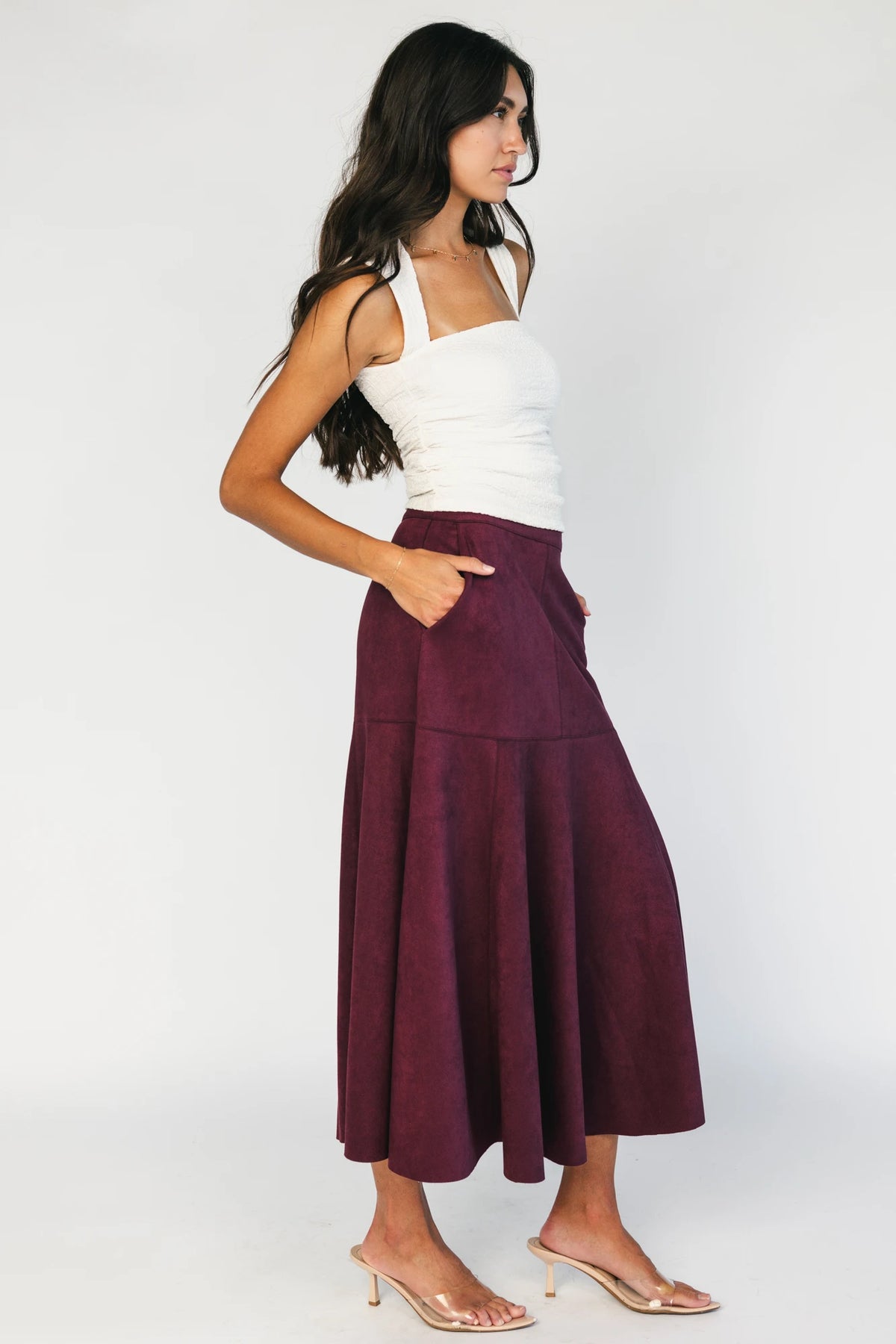 Model wearing a mulberry faux suede A-line midi skirt with a high-rise waistband, stitched panel detailing, side pockets, and a raw hem for a timeless, feminine silhouette.