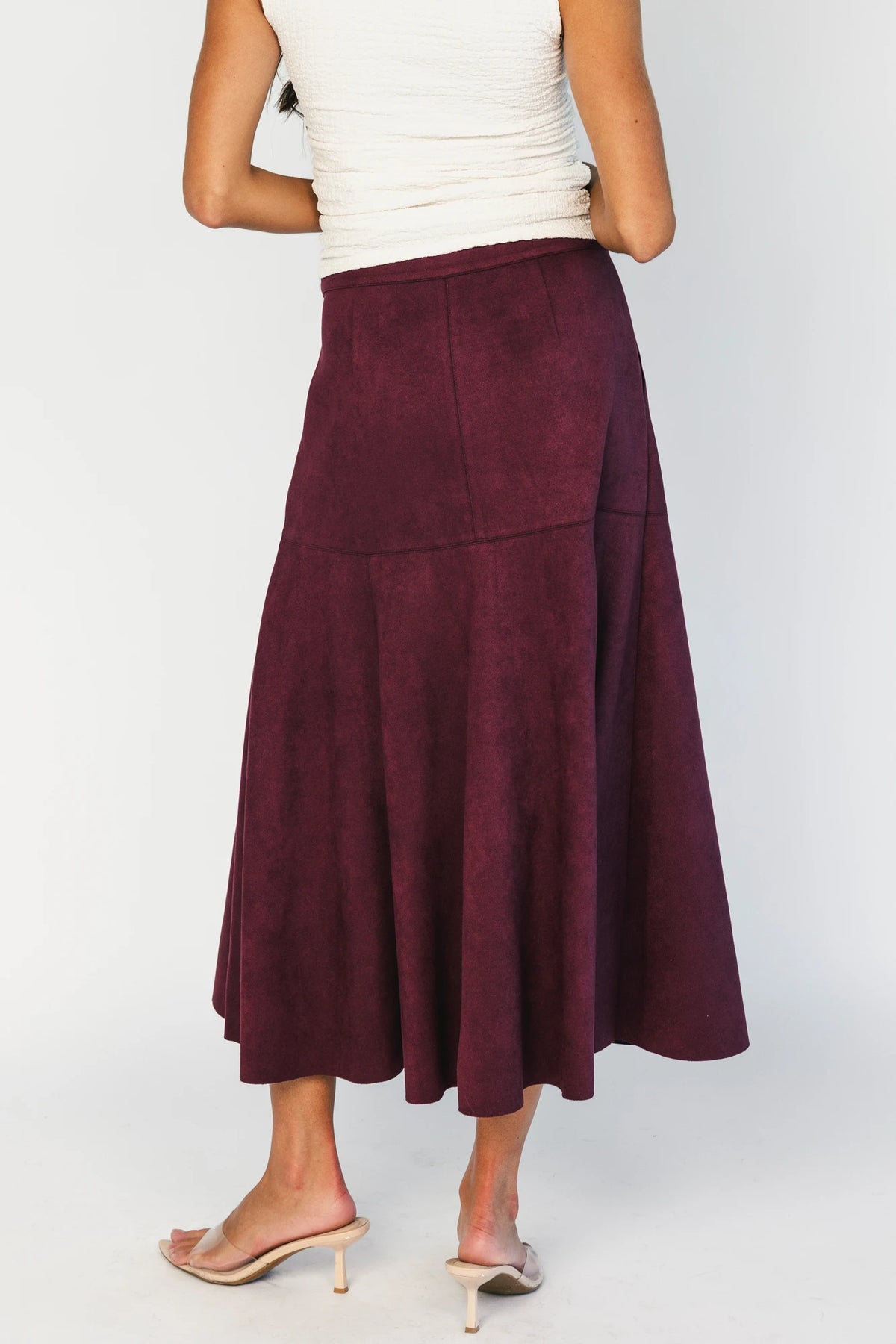 Model wearing a mulberry faux suede A-line midi skirt with a high-rise waistband, stitched panel detailing, side pockets, and a raw hem for a timeless, feminine silhouette.