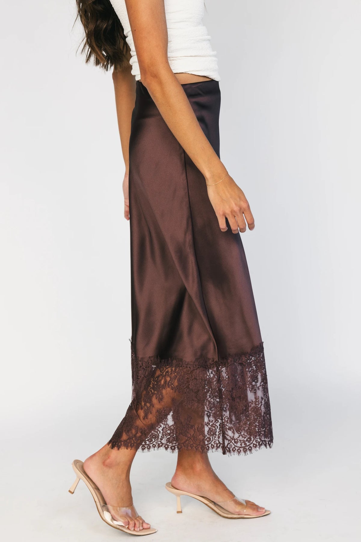 Model wearing the Kamara Satin Lace Skirt in Chocolate featuring a high-rise waistband, bias-cut satin silhouette, delicate floral lace trim at the hem, and a flowing midi length.
