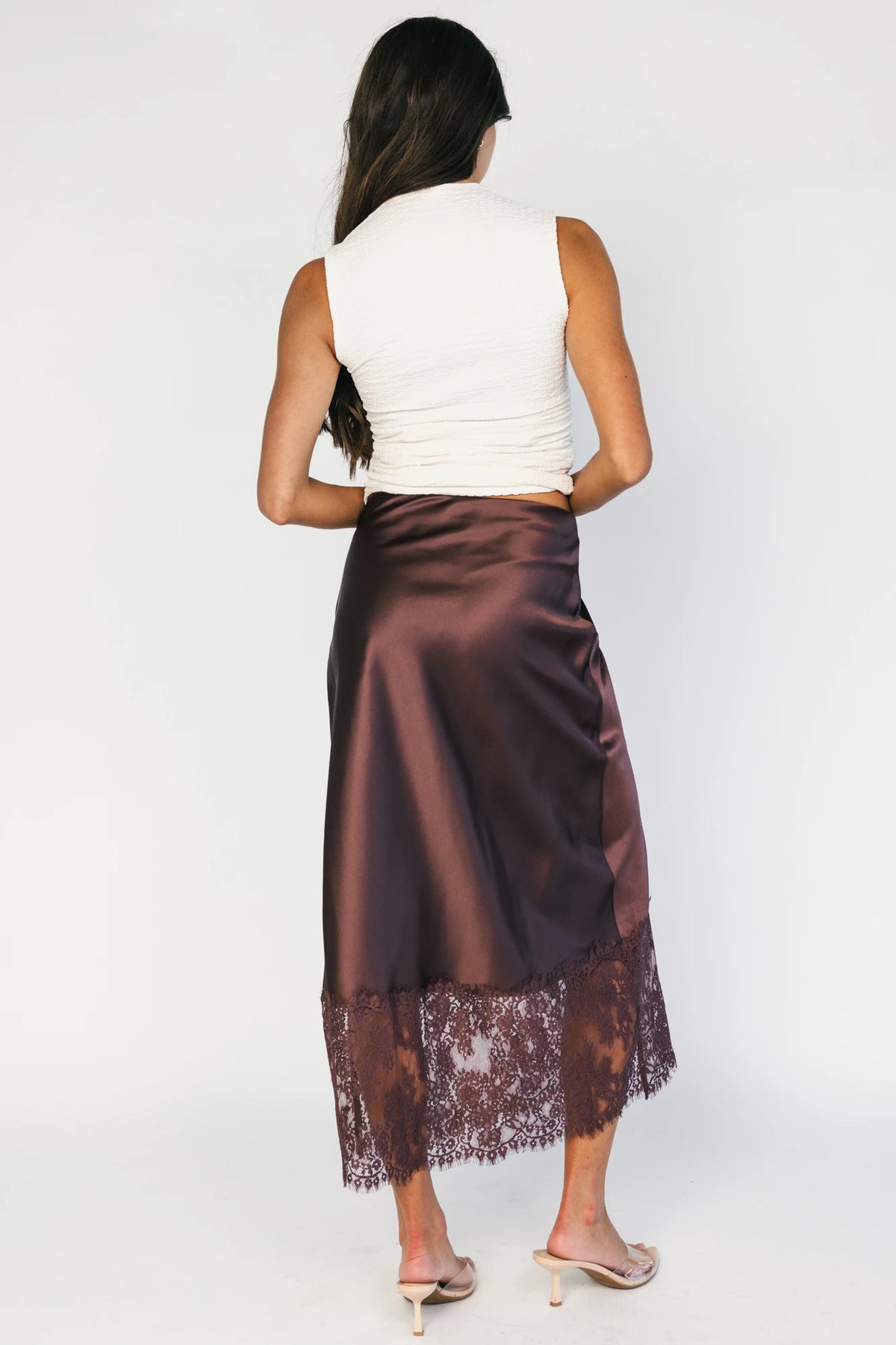 Model wearing the Kamara Satin Lace Skirt in Chocolate featuring a high-rise waistband, bias-cut satin silhouette, delicate floral lace trim at the hem, and a flowing midi length.