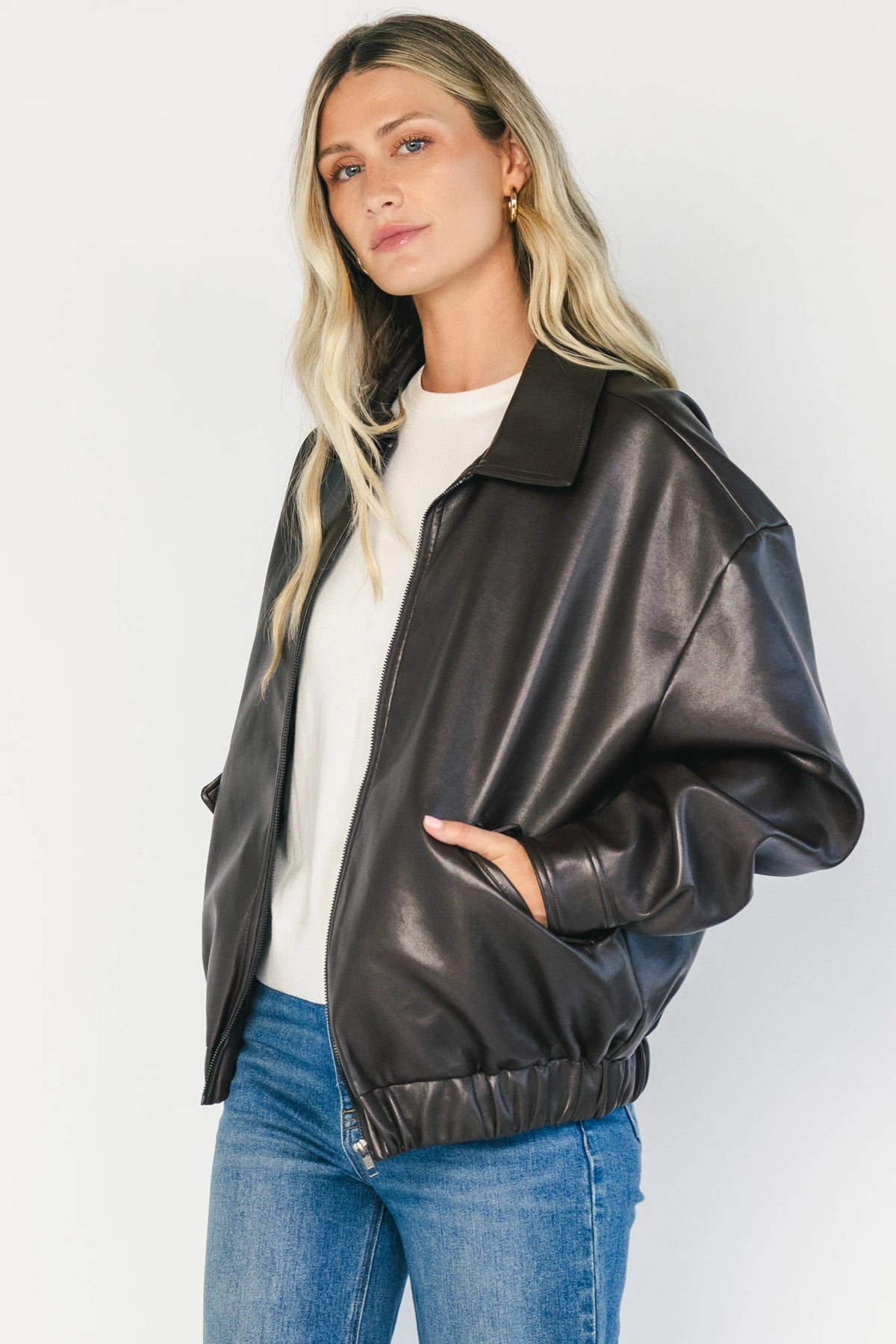 Model wearing a dark brown faux leather bomber jacket with a folded lapel collar, front zipper closure, elastic hem, silver snap cuffs, and front pockets.