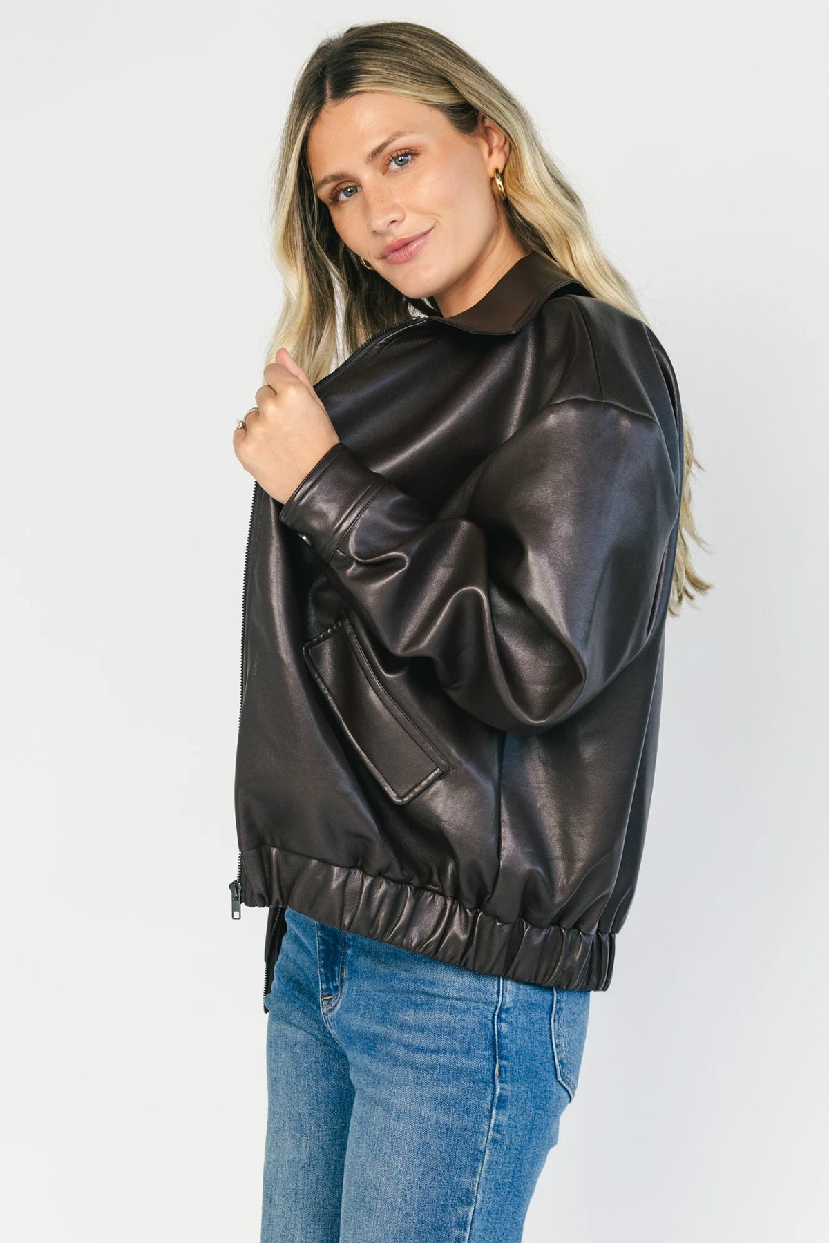 Model wearing a dark brown faux leather bomber jacket with a folded lapel collar, front zipper closure, elastic hem, silver snap cuffs, and front pockets.