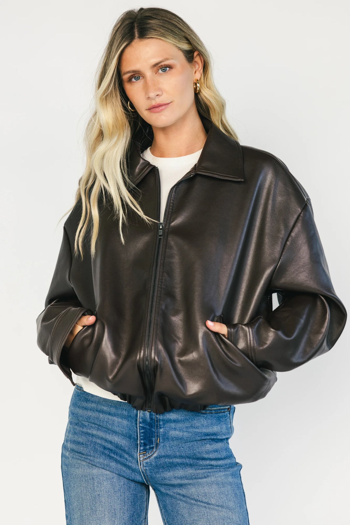 Model wearing a dark brown faux leather bomber jacket with a folded lapel collar, front zipper closure, elastic hem, silver snap cuffs, and front pockets.