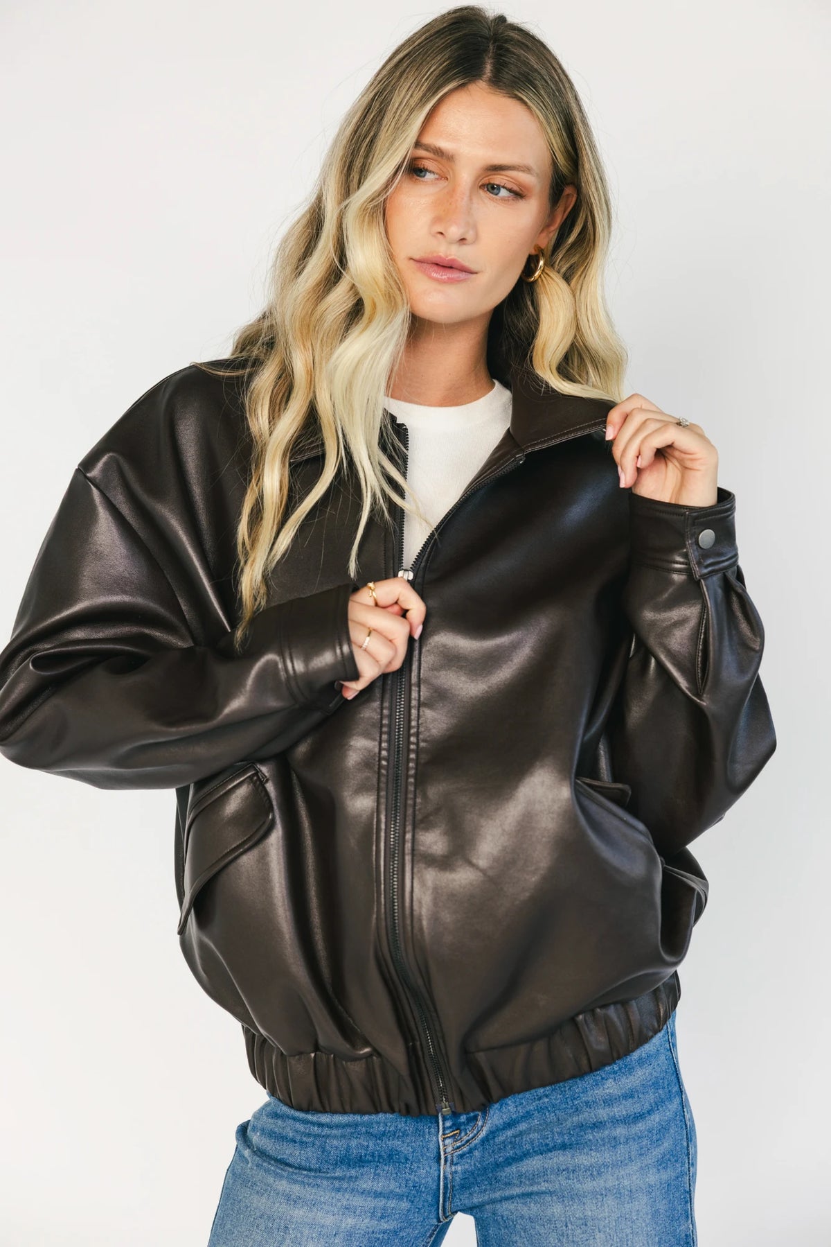 Model wearing a dark brown faux leather bomber jacket with a folded lapel collar, front zipper closure, elastic hem, silver snap cuffs, and front pockets.