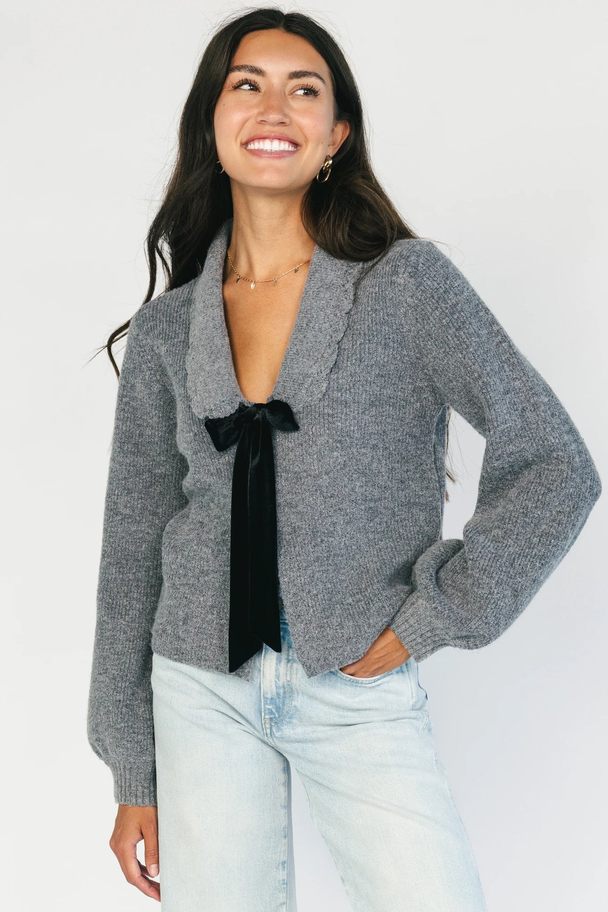 Model wearing a gray knit cardigan with a scalloped collar, adjustable black velvet bow detail, two front hook-and-eye closures, and a relaxed fit.