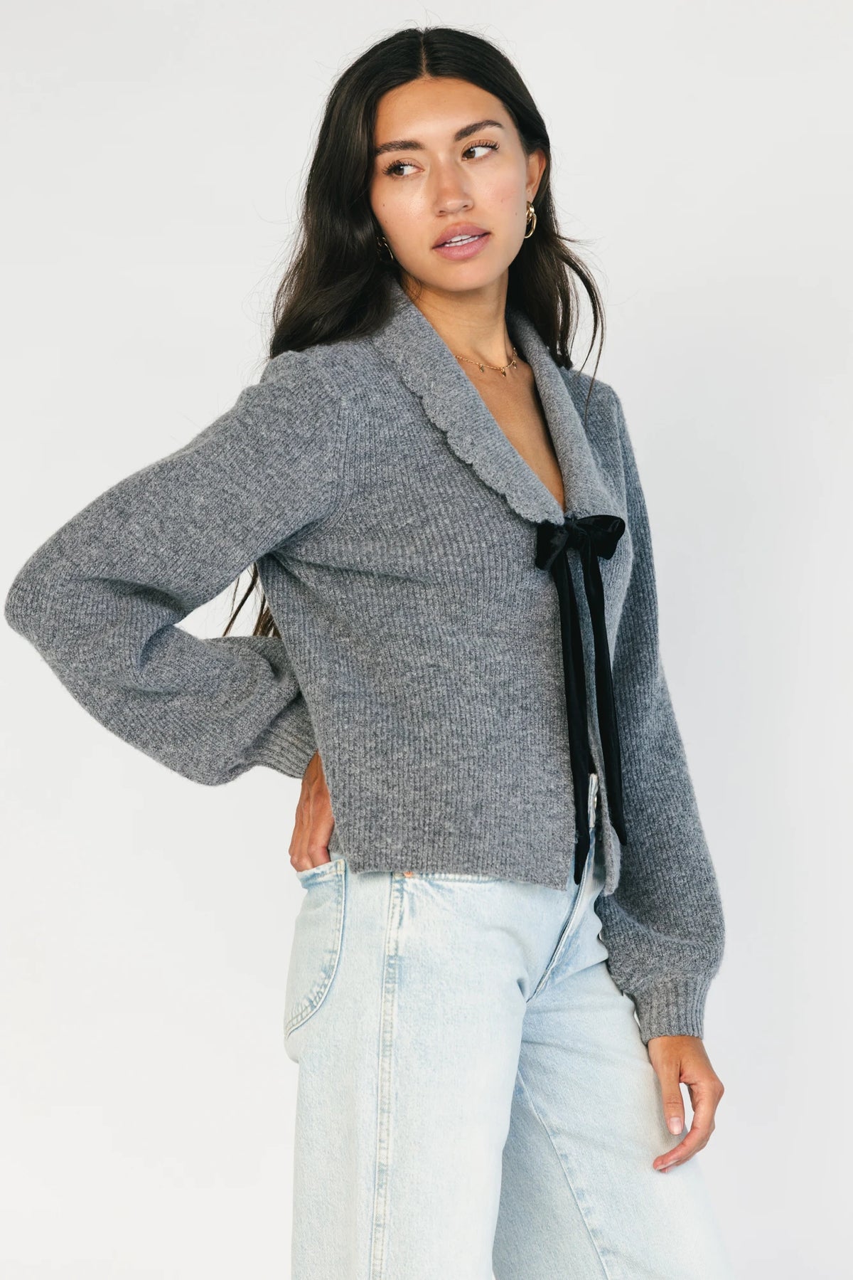 Model wearing a gray knit cardigan with a scalloped collar, adjustable black velvet bow detail, two front hook-and-eye closures, and a relaxed fit.