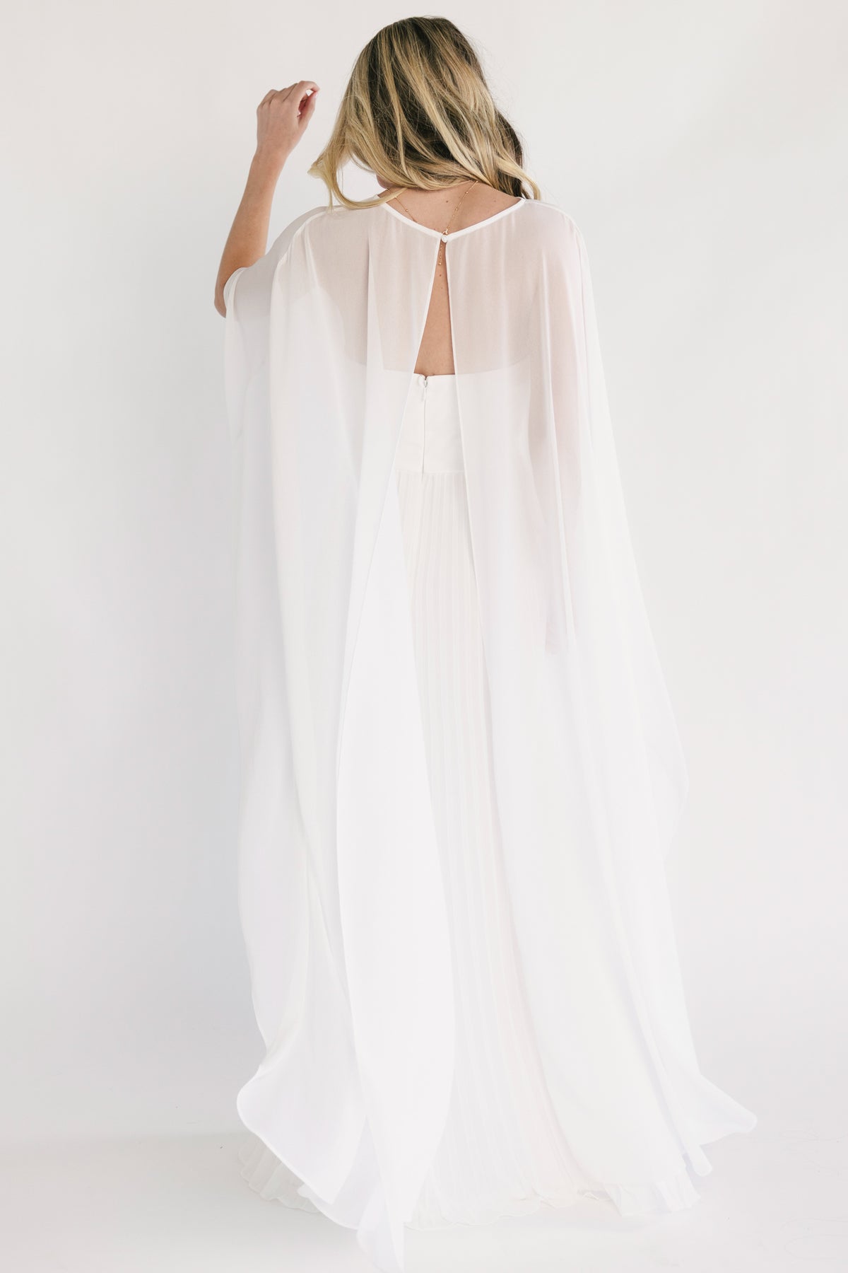 Three-quarter view of gown highlighting pleated skirt movement and soft chiffon cape drape over the arms.