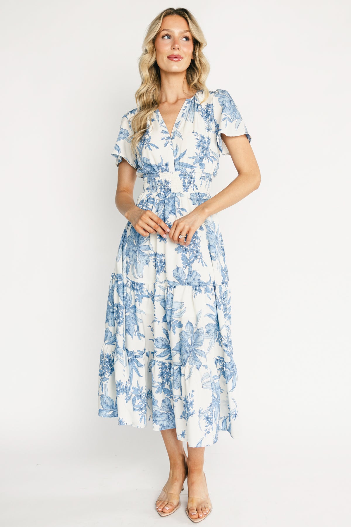 Front view of a model wearing the Holland smocked midi dress in off-white with blue floral print, V-neck, smocked waist, and flutter sleeves.