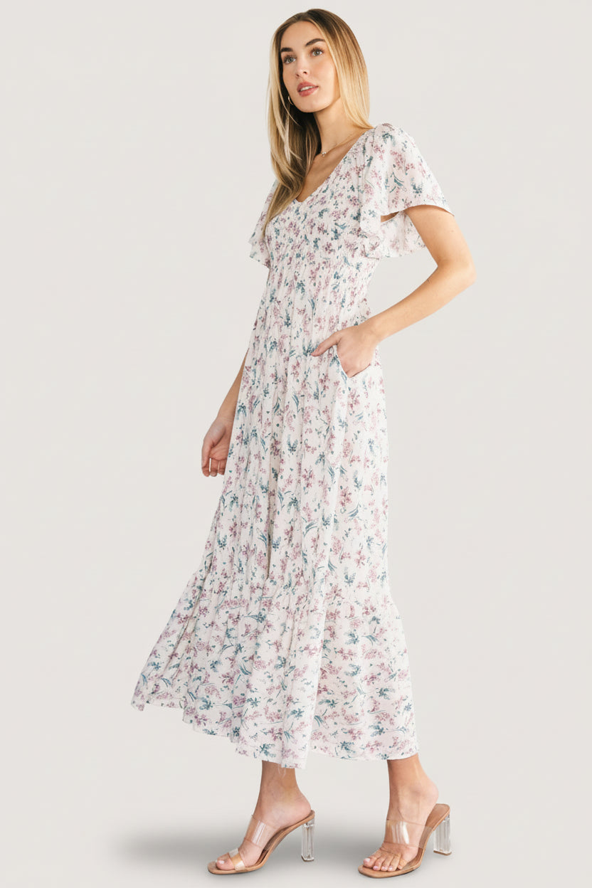 Model wearing the Joanie smocked midi dress in cream multi floral with hands in hidden pockets, showcasing relaxed silhouette and flutter sleeves.