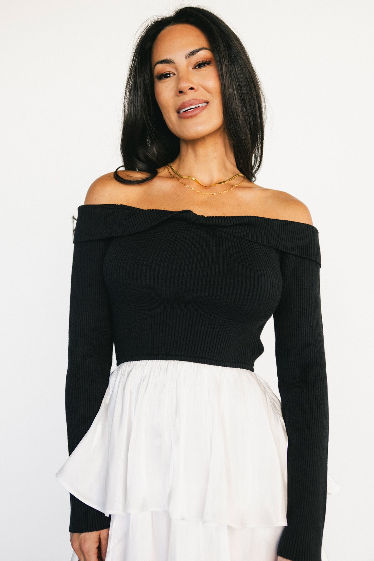 Three-quarter view showing elastic twist off-shoulder neckline and long sweater sleeves paired with white satin tiered ruffle skirt.