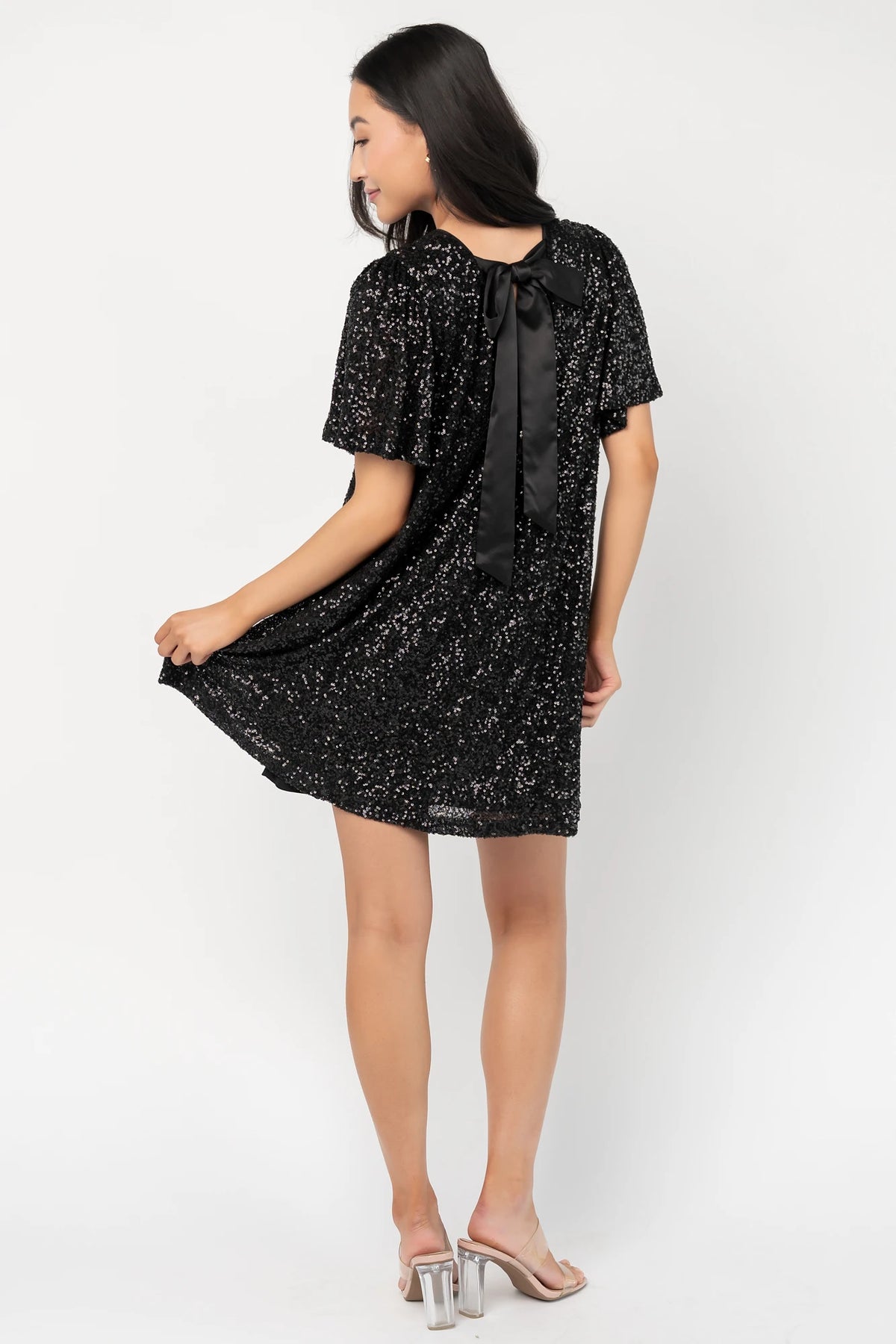 Black liquid sequin mini shift dress with a round neckline, short flutter sleeves, and a satin bow back detail.