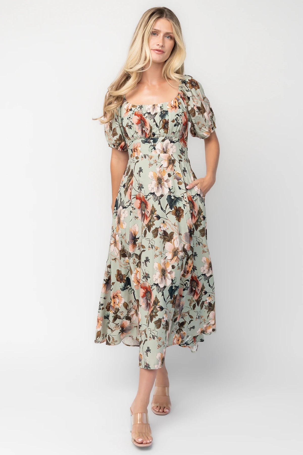 Model wearing the Charlotte Midi Dress in Sage, a sage green midi dress with a pink multi floral print, rounded square neckline, puff sleeves, banded waist, and flowing A-line skirt with side pockets.