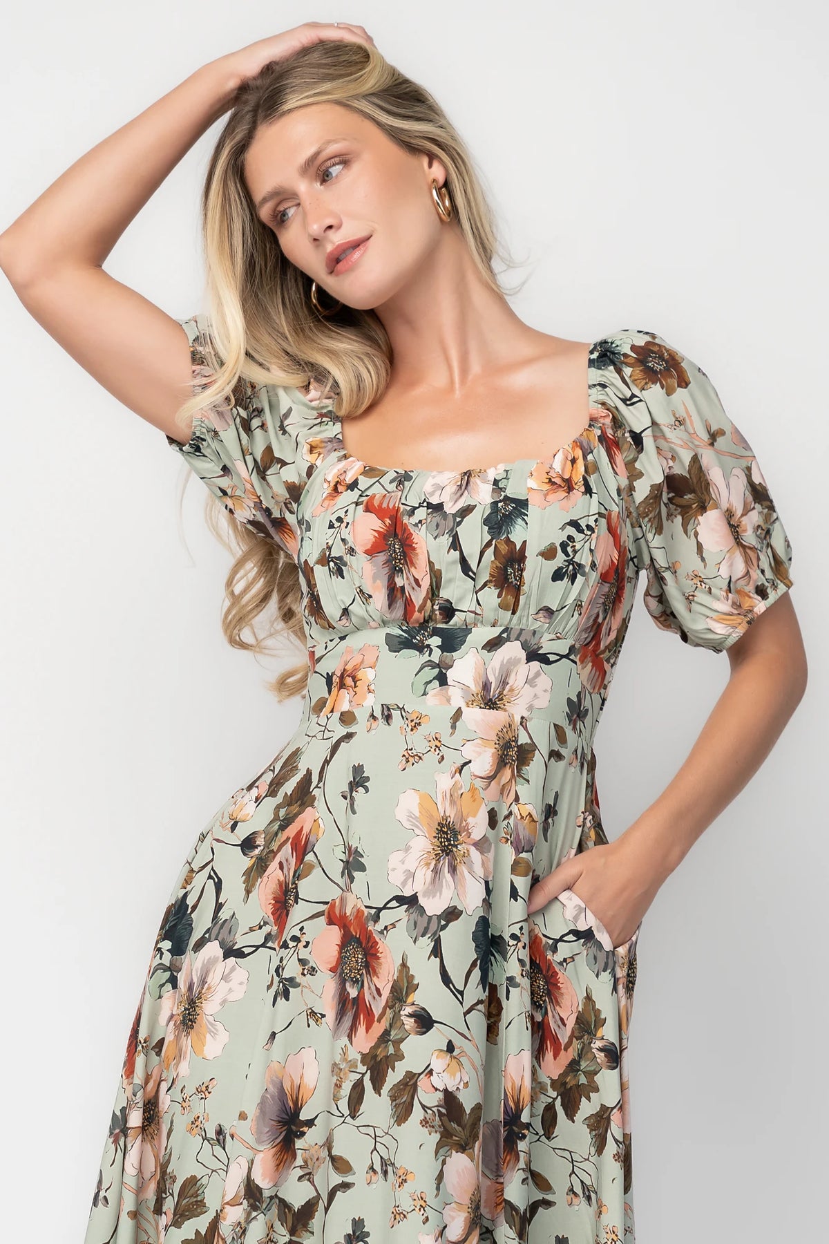 Model wearing the Charlotte Midi Dress in Sage, a sage green midi dress with a pink multi floral print, rounded square neckline, puff sleeves, banded waist, and flowing A-line skirt with side pockets.