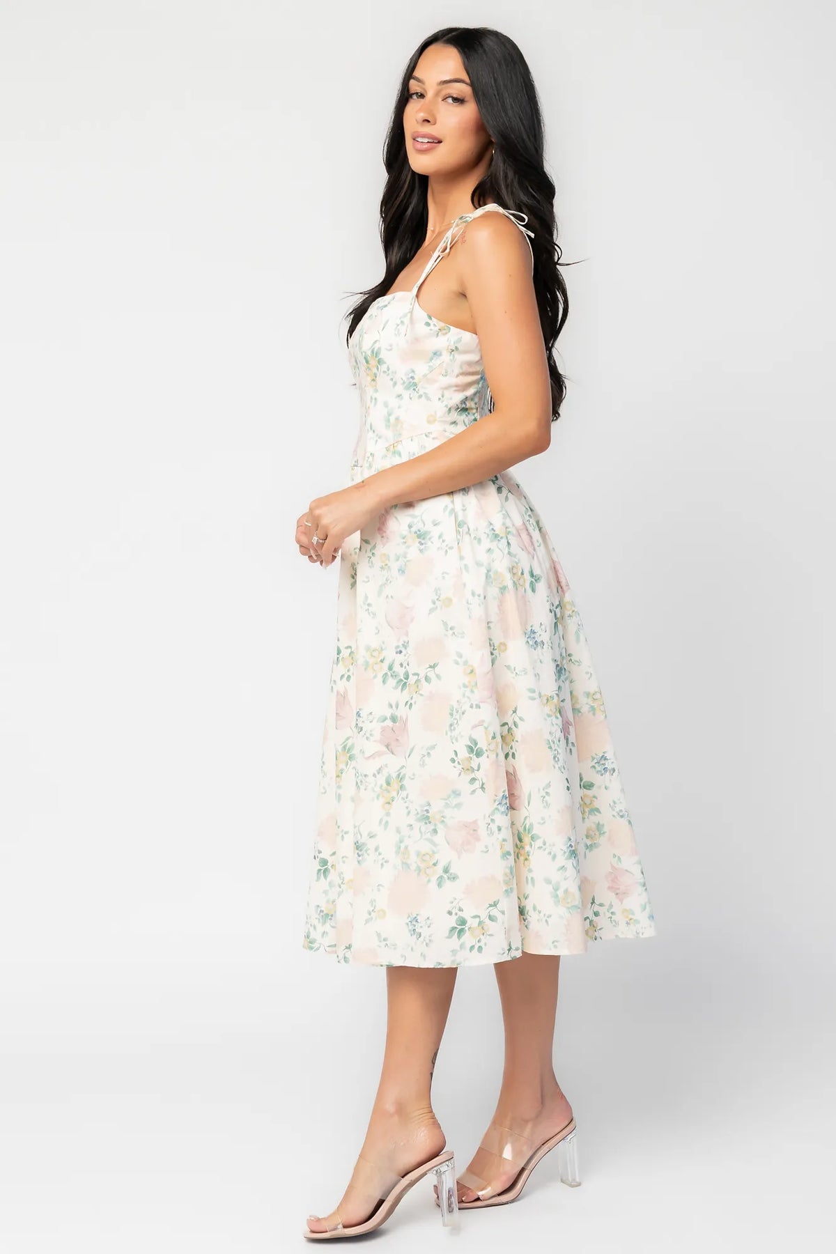 Side image of model wearing a cream floral midi dress with a square neckline, adjustable shoulder tie straps, fitted bodice, and flowing skirt. 
