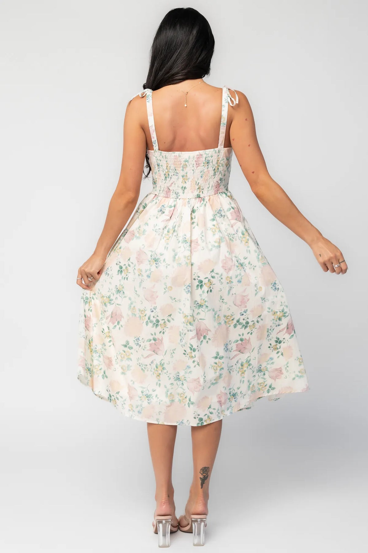 Back image of model wearing a cream floral midi dress with a square neckline, adjustable shoulder tie straps, fitted bodice, and flowing skirt. 