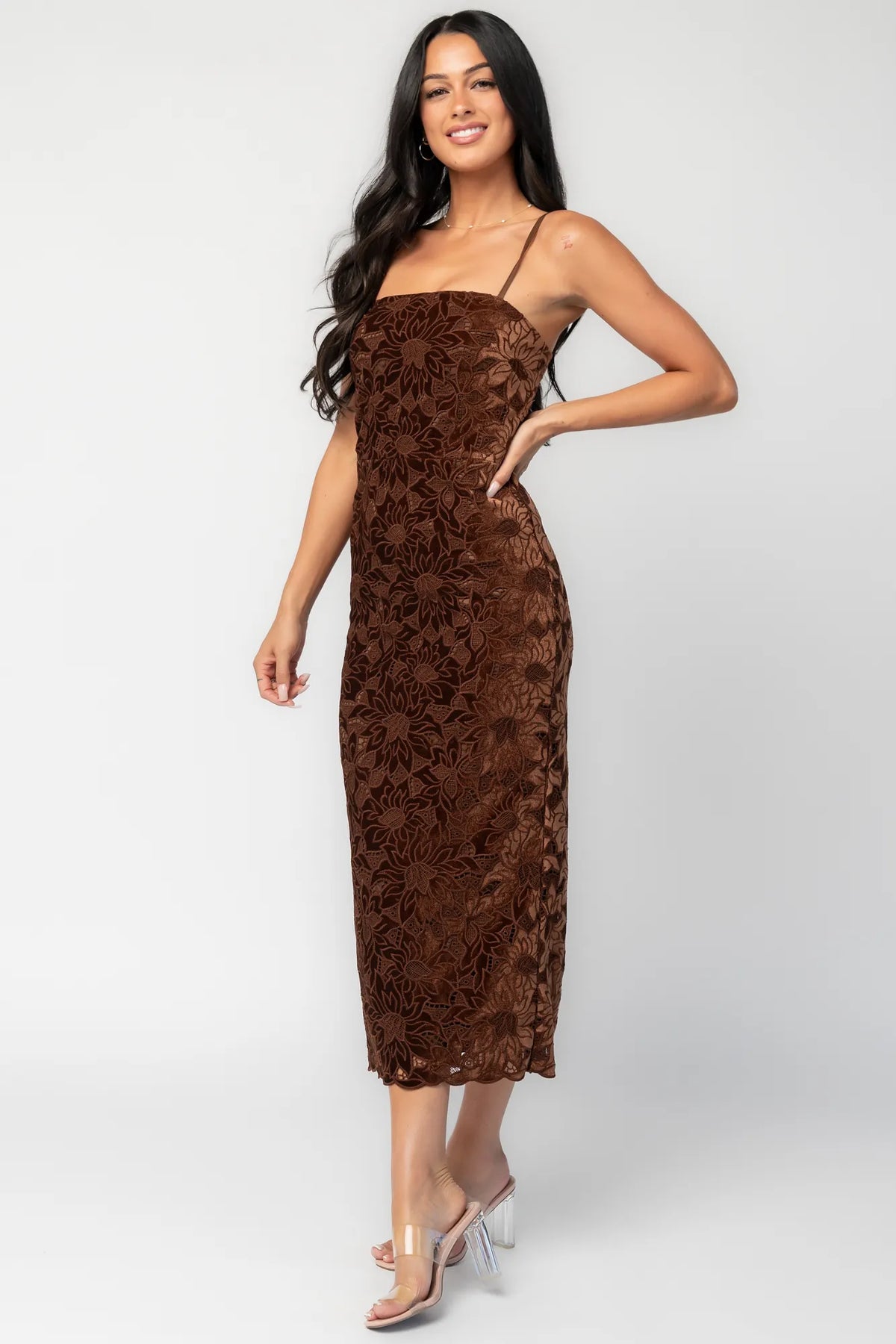 Model wearing a fitted espresso brown midi dress featuring an allover floral velvet lace overlay. The dress has a square neckline, adjustable spaghetti straps, and a body-skimming silhouette that falls below the knee with a scalloped hem.